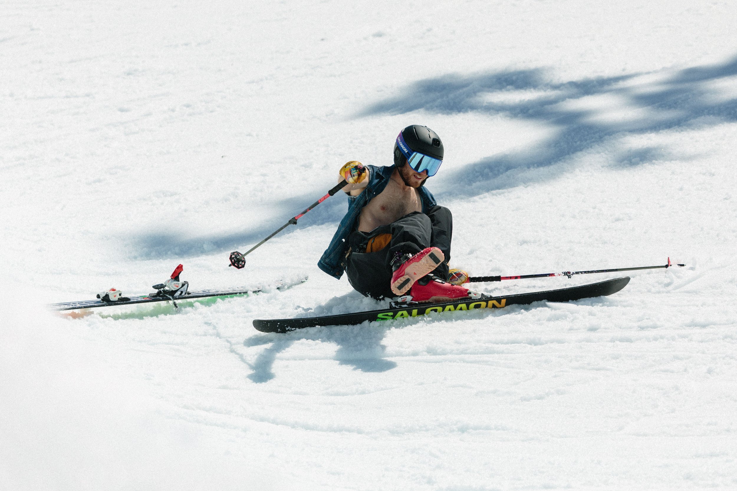 A man has fallen off his skis on a snow-covered slope, wearing a helmet and goggles, with his shirt unbuttoned, and skis and poles nearby.