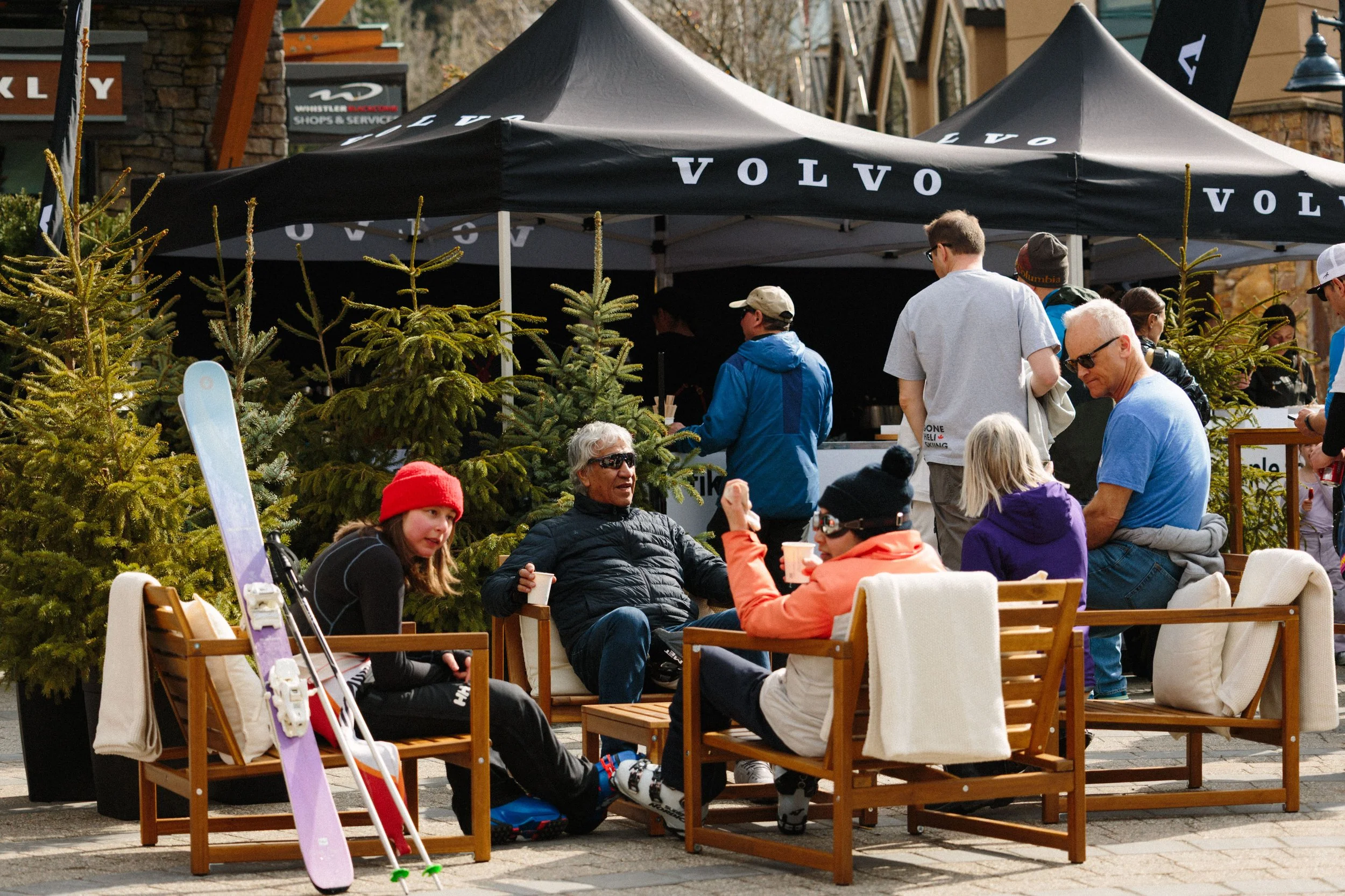 People sitting on chairs and standing under a black canopy with the Volvo logo, at an outdoor event with pine trees and buildings in the background.