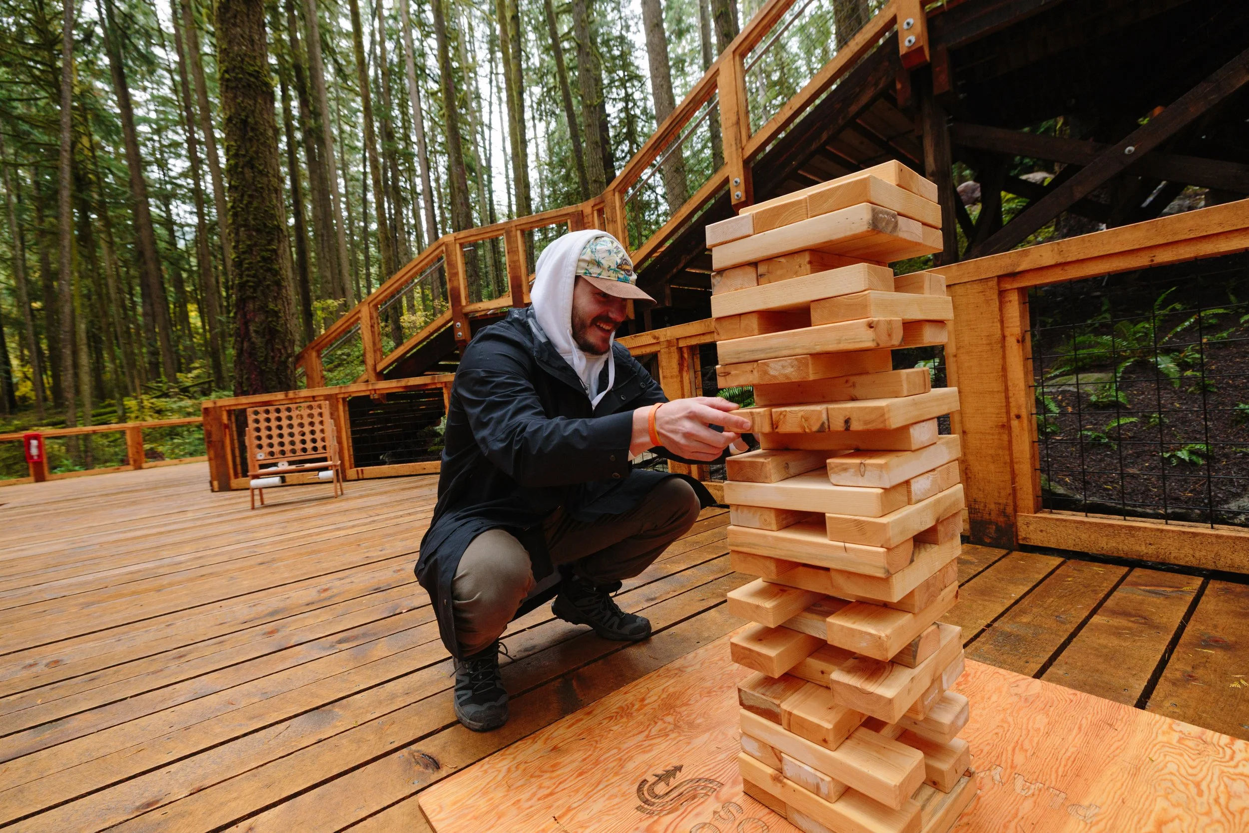 A man playing giant Jenga outdoors on a wooden deck surrounded by trees.