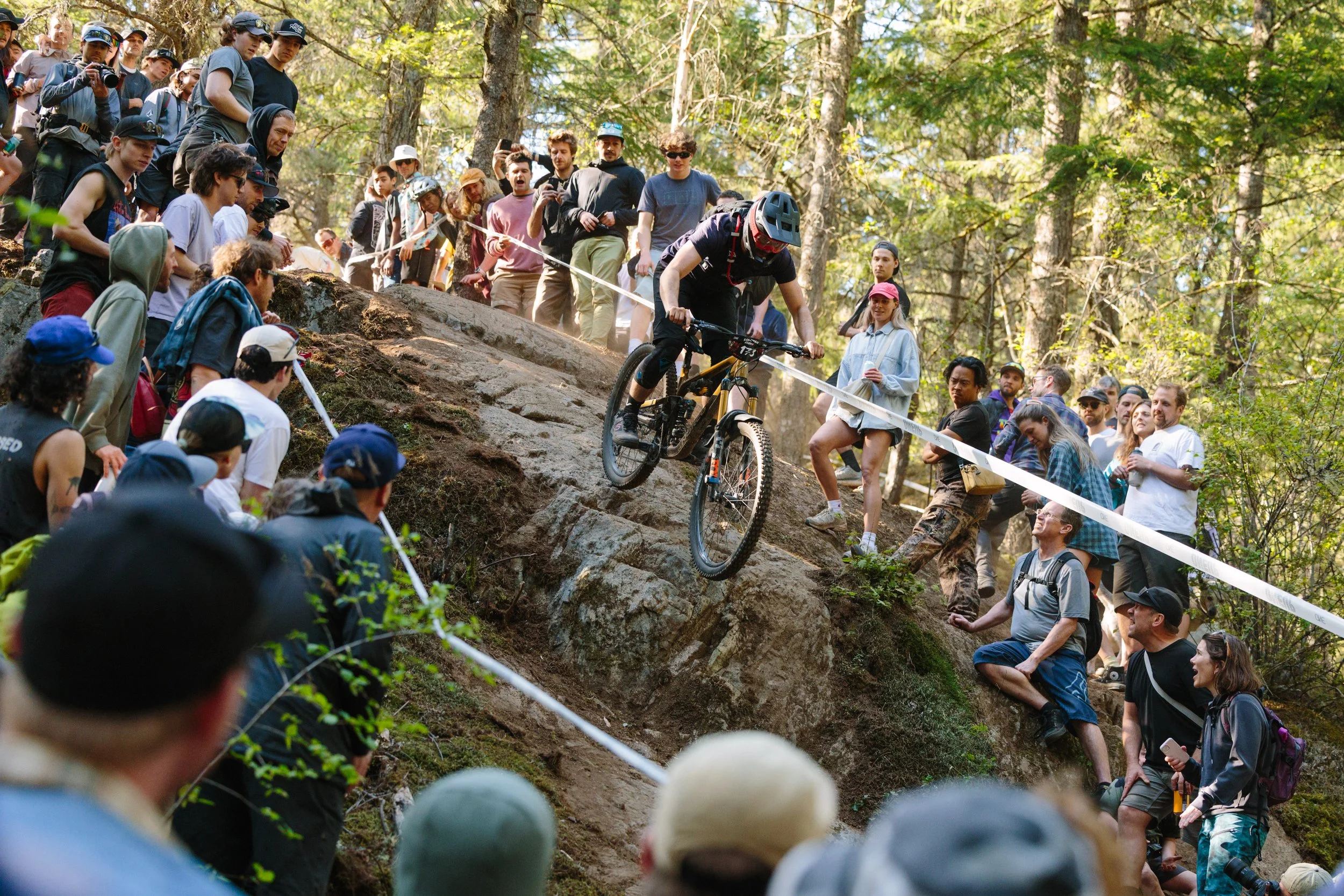 A mountain biker in a helmet and black clothing descending a rocky trail on a steep slope, surrounded by a large crowd of spectators and fellow riders in a forest setting.