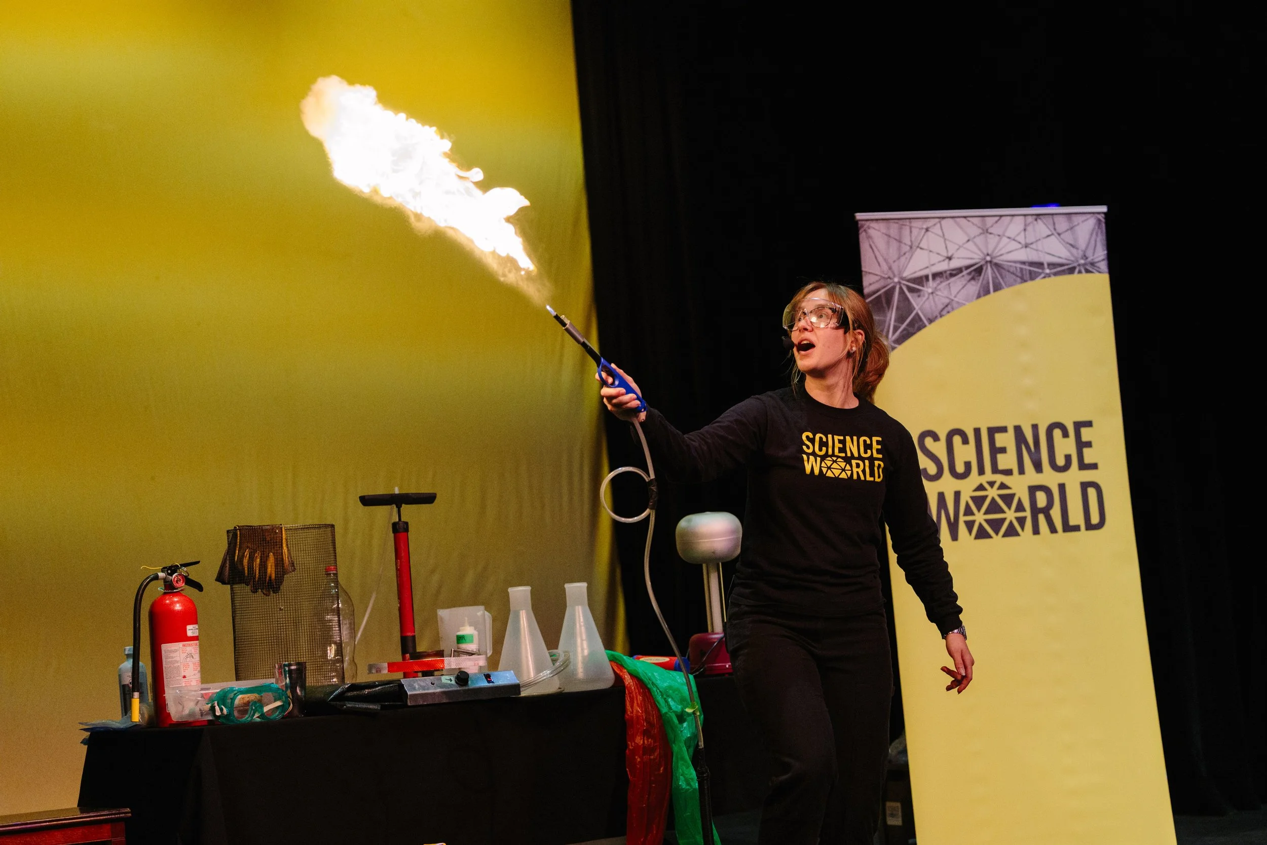 A woman in black Science World shirt is performing a science demonstration involving fire and gas on stage, with scientific equipment and a Science World banner in the background.