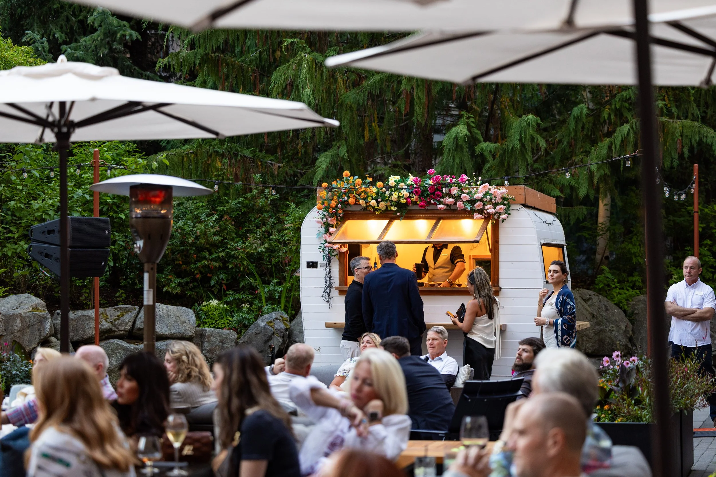 People at an outdoor event gathered around a white food truck decorated with flowers, with trees and large rocks in the background.