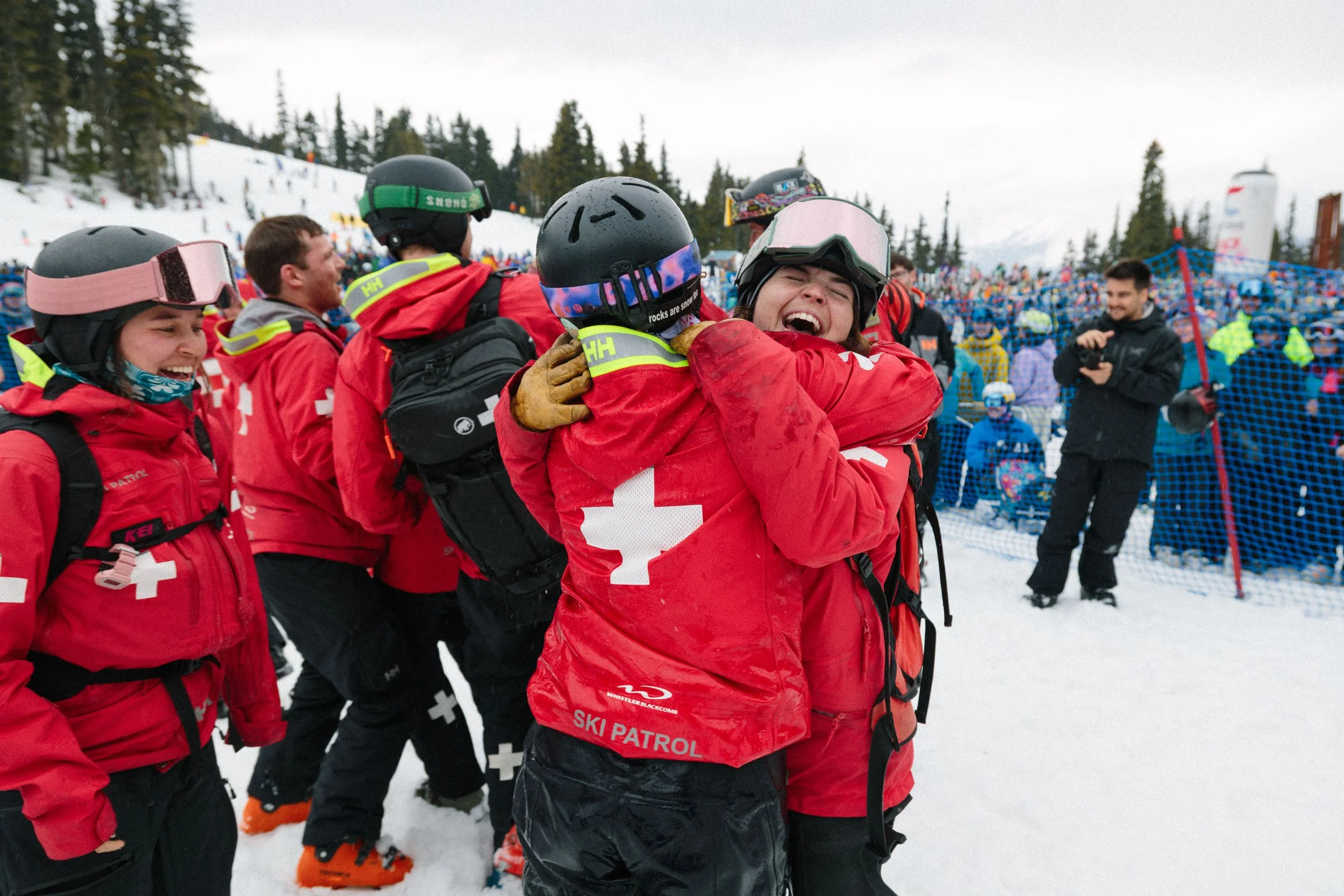 A group of people in red jackets, some with helmets, are celebrating and hugging on a snowy mountain with others in blue jackets in the background.