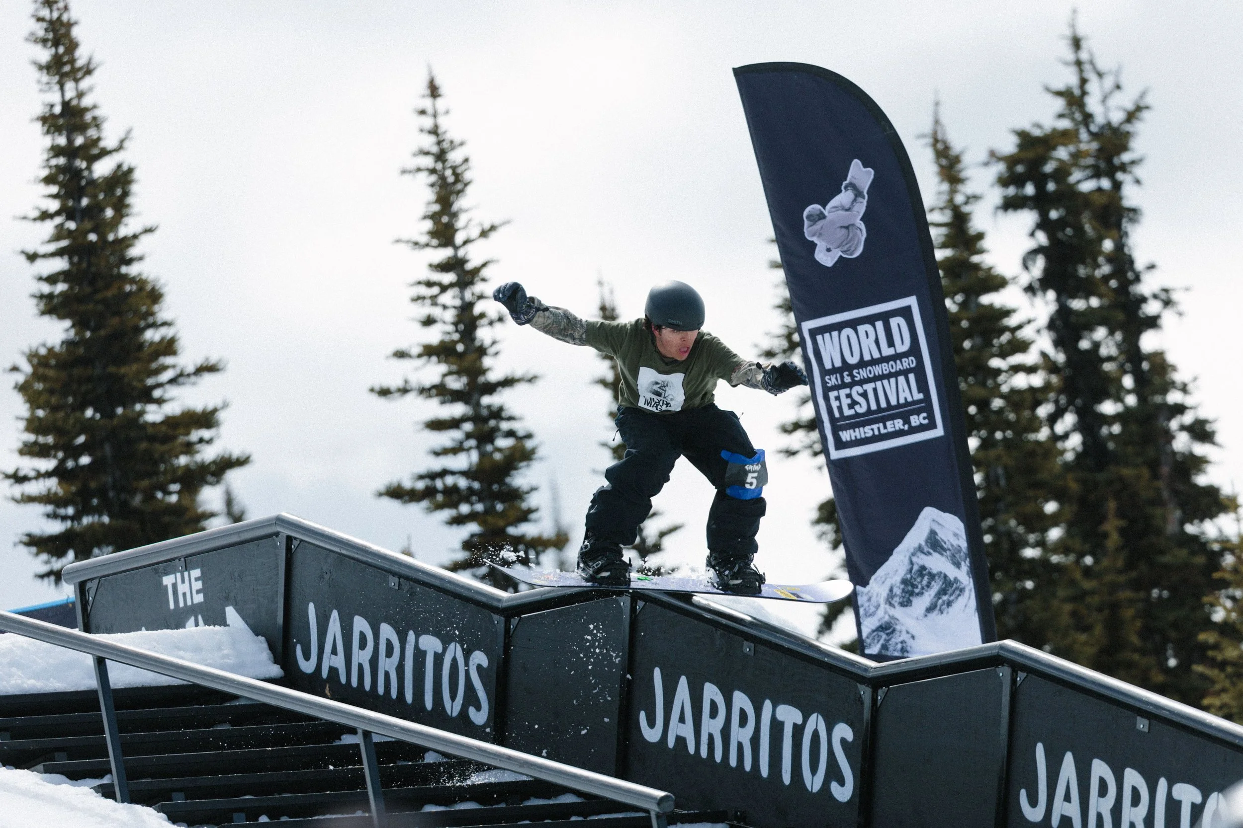 A snowboarder in a helmet and green shirt riding on a skate ramp at an outdoor snow park during a snowboarding event, with trees in the background and a banner that reads 'JARRITOS' and 'WORLD SKI & SNOWBOARD FESTIVAL WHISTLER, BC'.
