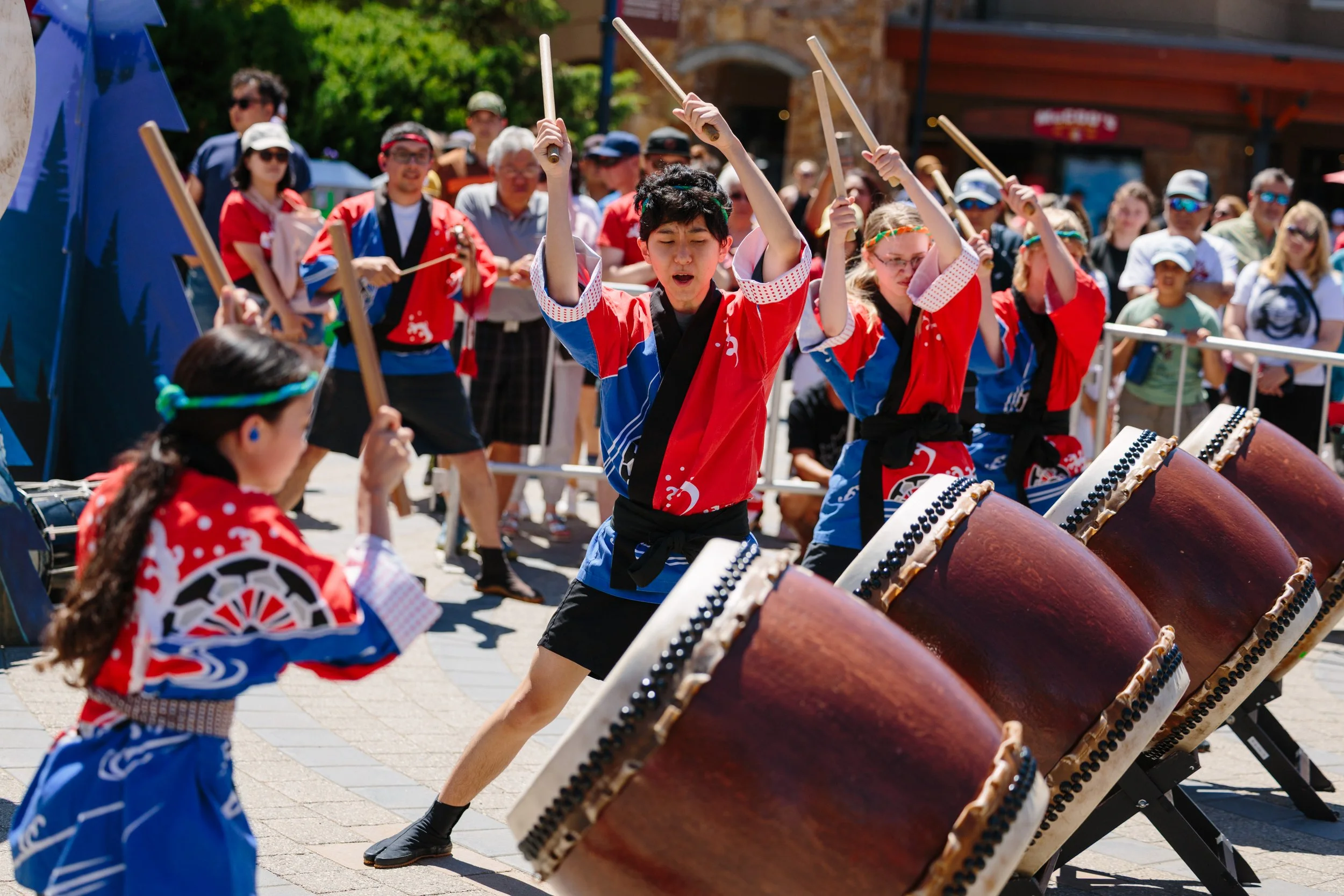 Children dressed in traditional red, blue, and black Japanese attire perform taiko drumming at an outdoor festival, surrounded by a crowd of observers.