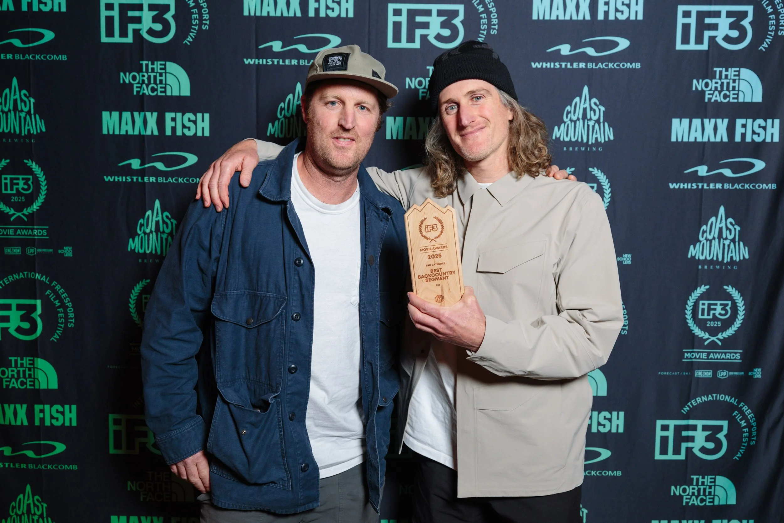 Two men standing together at an awards event, one holding a wooden trophy, posing in front of a backdrop with logos of IF3, The North Face, Coast Mountain Brewing, and Whistler Blackcomb, smiling.
