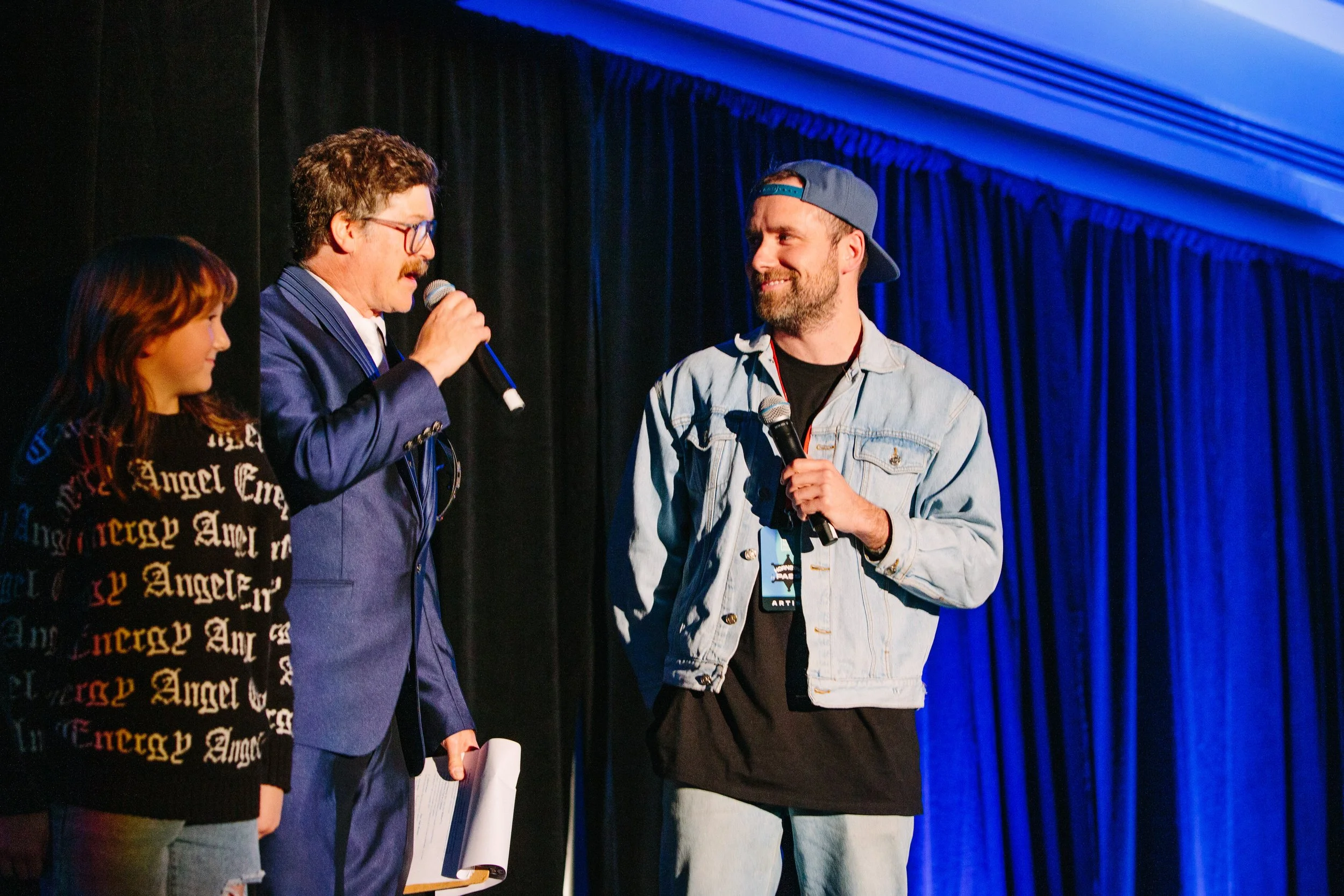 Three people on stage with black curtains and blue lighting. A woman on the left looks at two men holding microphones. The man in the middle, wearing glasses and a navy suit, speaks into his microphone. The man on the right, wearing a backward cap, d