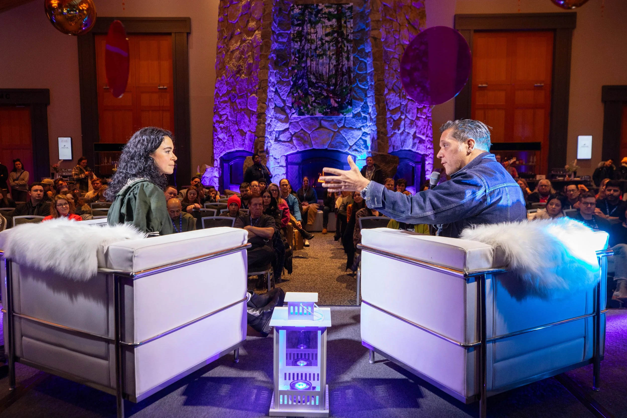 A woman and a man are seated in armchairs on a stage, having a conversation or interview in front of an audience in a large, decorated room with a fireplace and purple lighting. The man is gesturing with his hand while speaking, and the woman is list