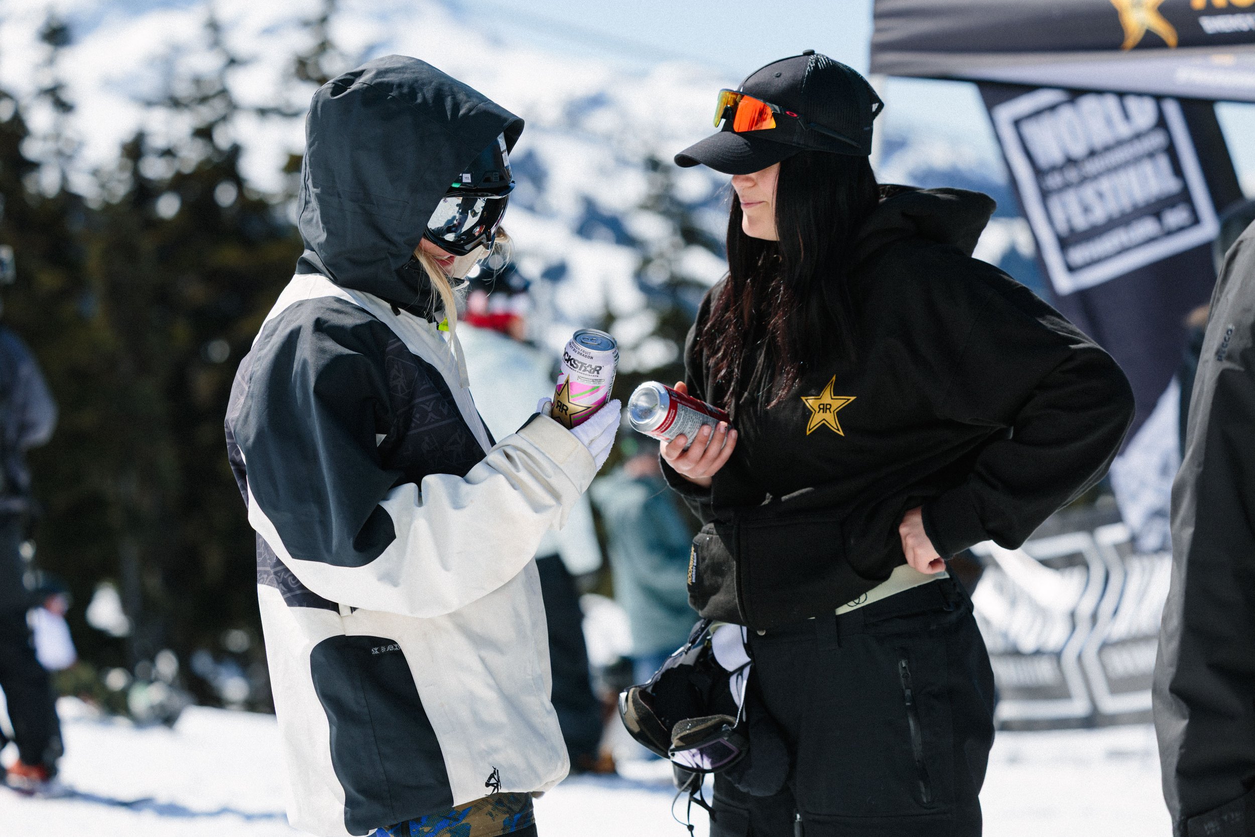 Two women in winter clothing and ski gear are having a conversation outdoors in a snowy area, with trees and a tent visible in the background.