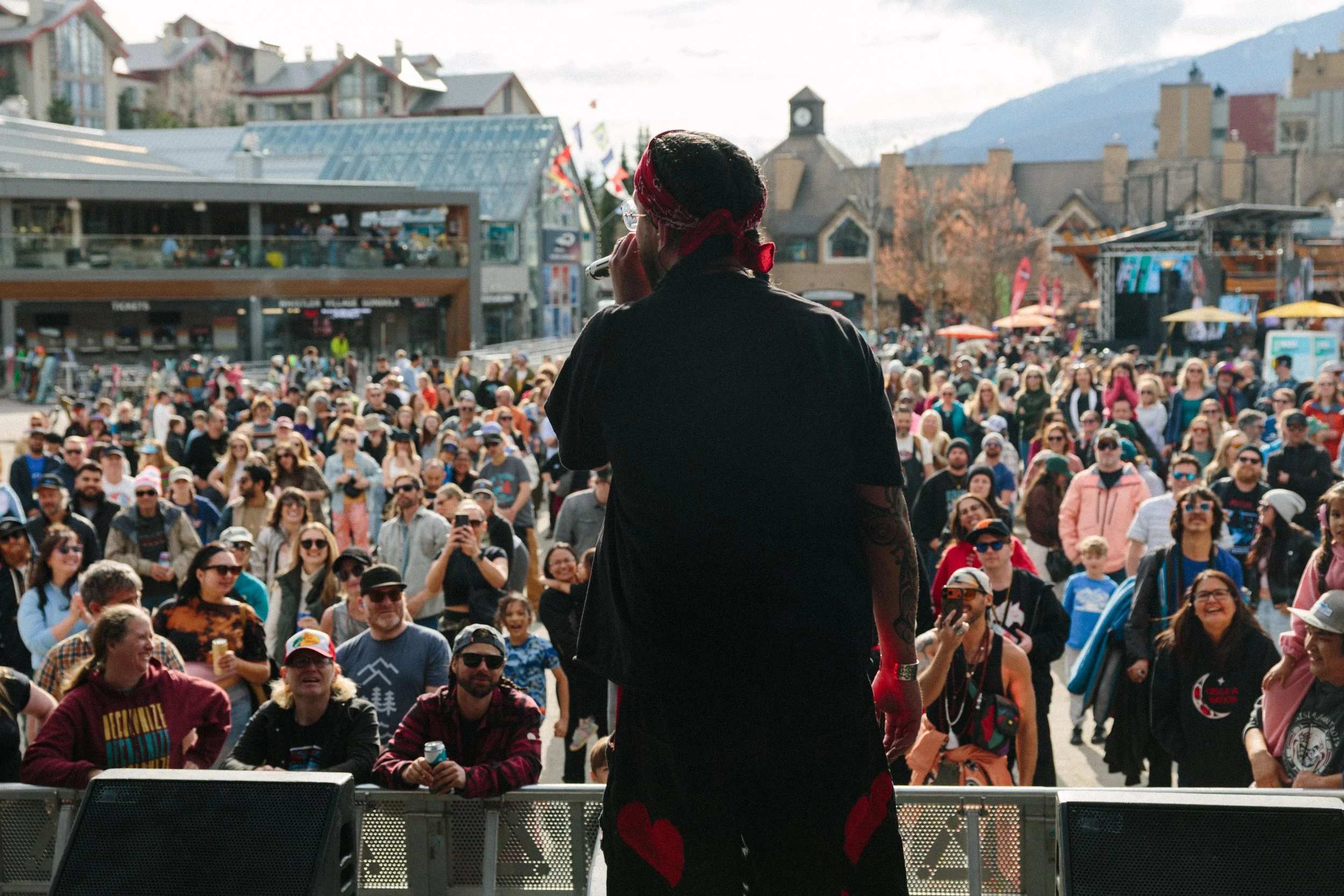 Performer on stage with crowd in the background, mountains visible in the distance, at an outdoor event.