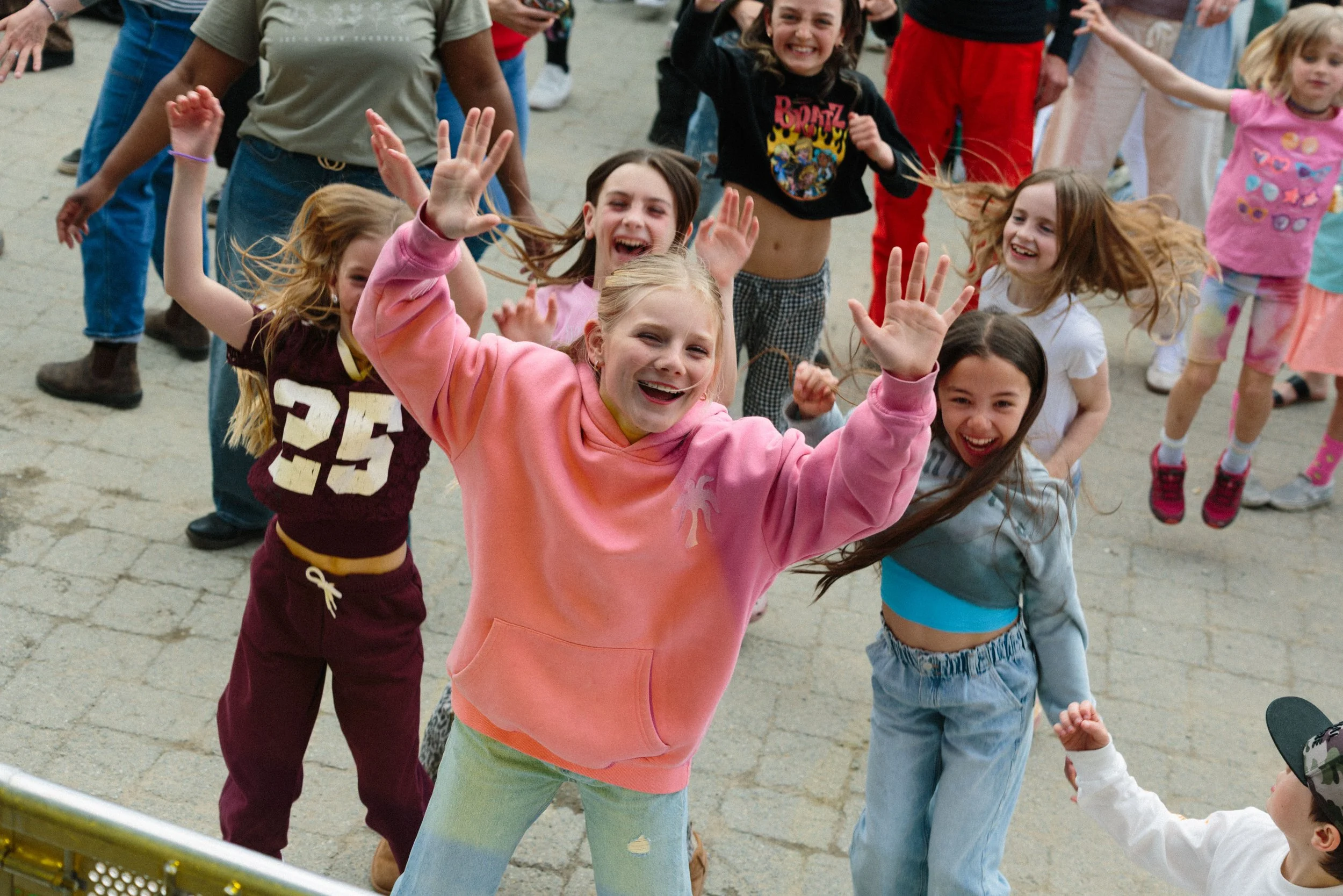 Group of children happily dancing and raising their hands outdoors on a paved area, with some adults visible in the background.