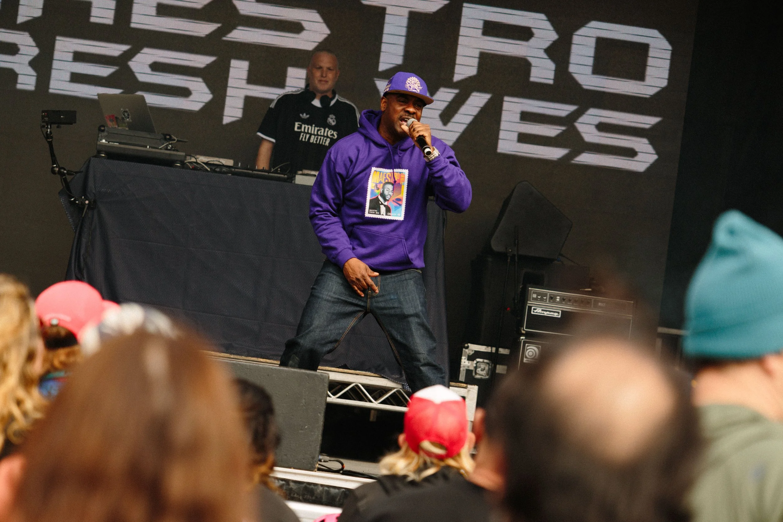 A man wearing a purple hoodie and matching baseball cap, holding a microphone and performing on stage. A DJ with a laptop and equipment stands behind him. The background features a large digital display with geometric patterns. Audience members are v