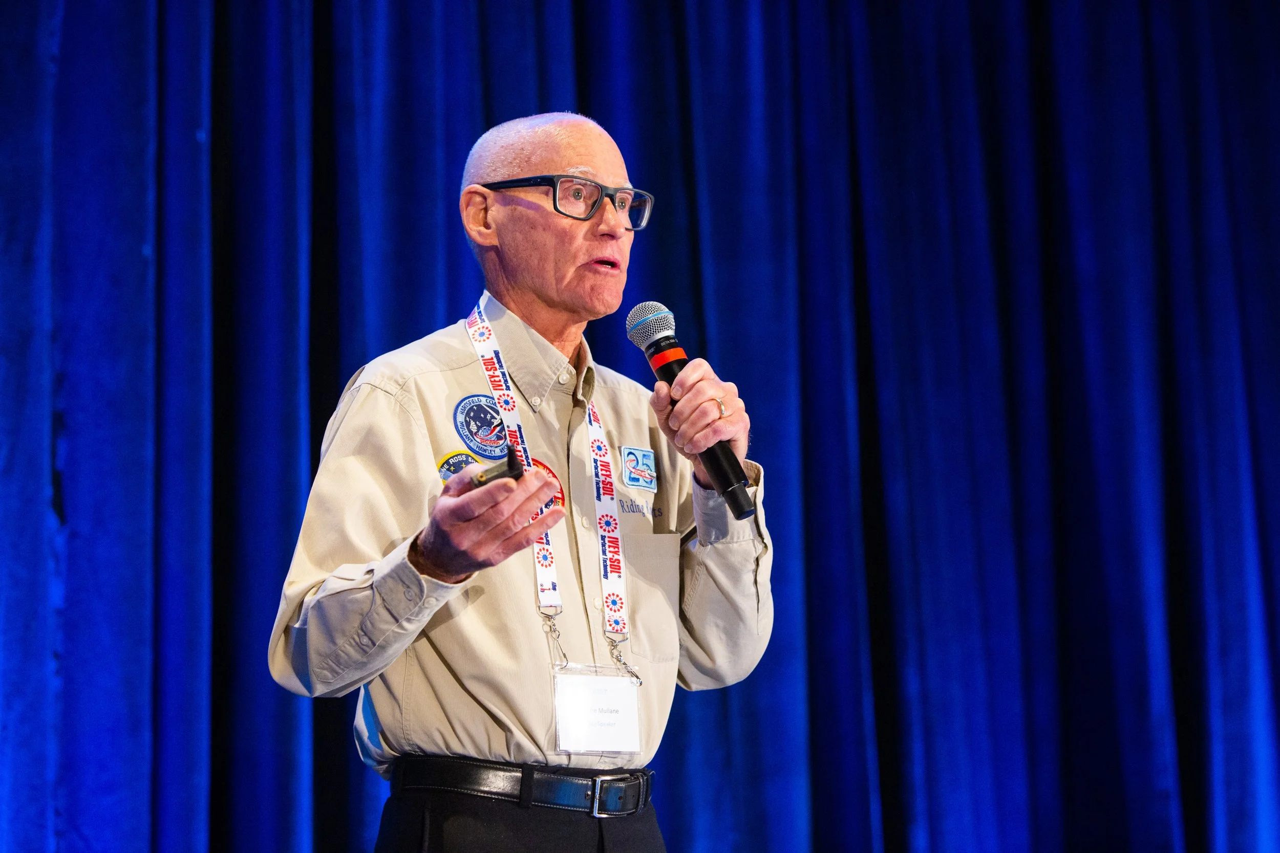 Speaker holding microphone and wearing conference lanyard against blue curtain backdrop.