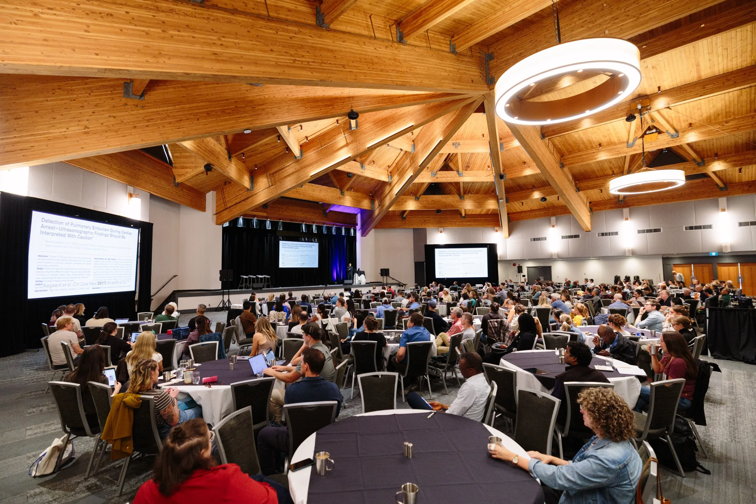 A large conference room filled with people attending a presentation with multiple screens displaying slides. The room has a wooden ceiling with modern circular light fixtures.