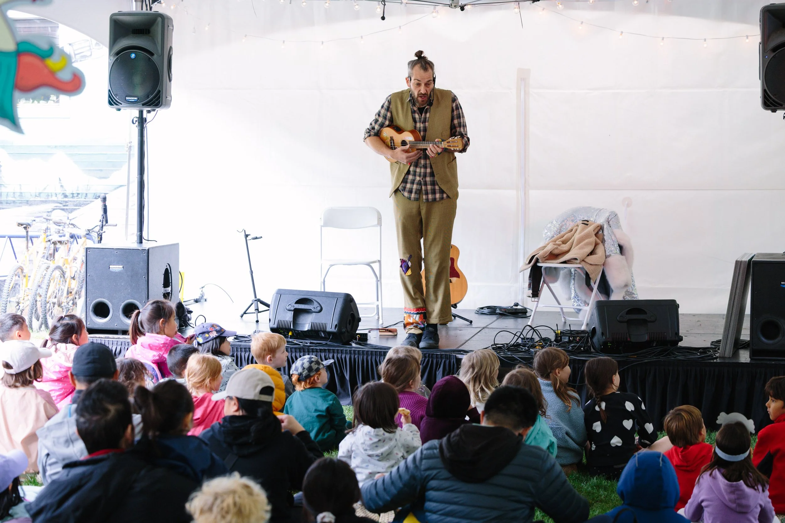 An adult performer playing a ukulele on stage at a children's outdoor event with kids sitting on grass watching.