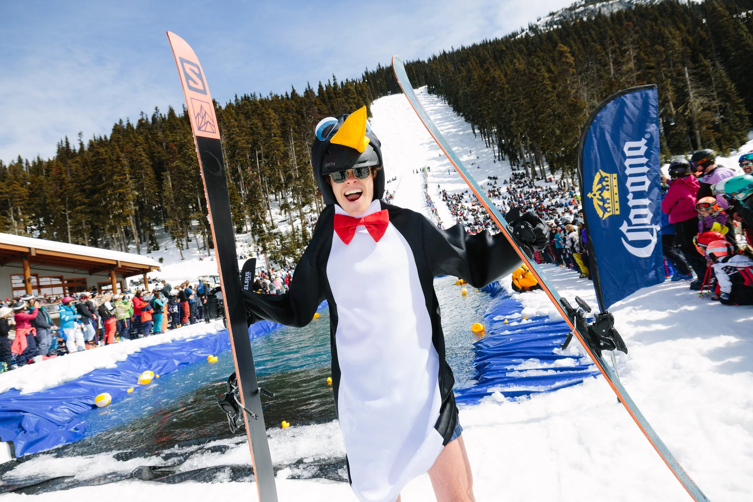 Person dressed as a penguin celebrating after a swimming event at a snowy outdoor winter competition, with a crowd of spectators and a ski slope in the background.