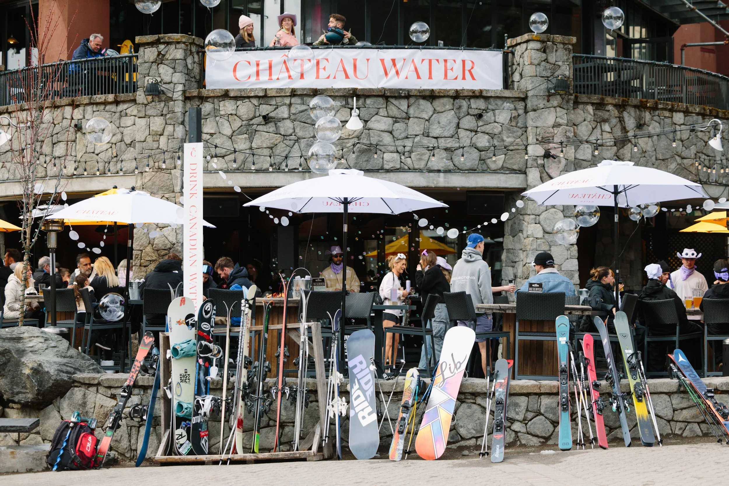 Outdoor patio of a restaurant or bar with people sitting at tables under large umbrellas, with a stone building in the background. Ski and snowboard equipment is displayed in front of the patio.