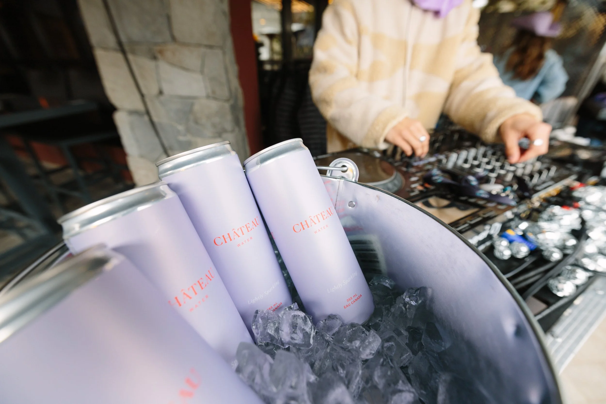 Four cans of Château Water in ice bucket, with a DJ wearing a beige fleece jacket mixing music in the background.