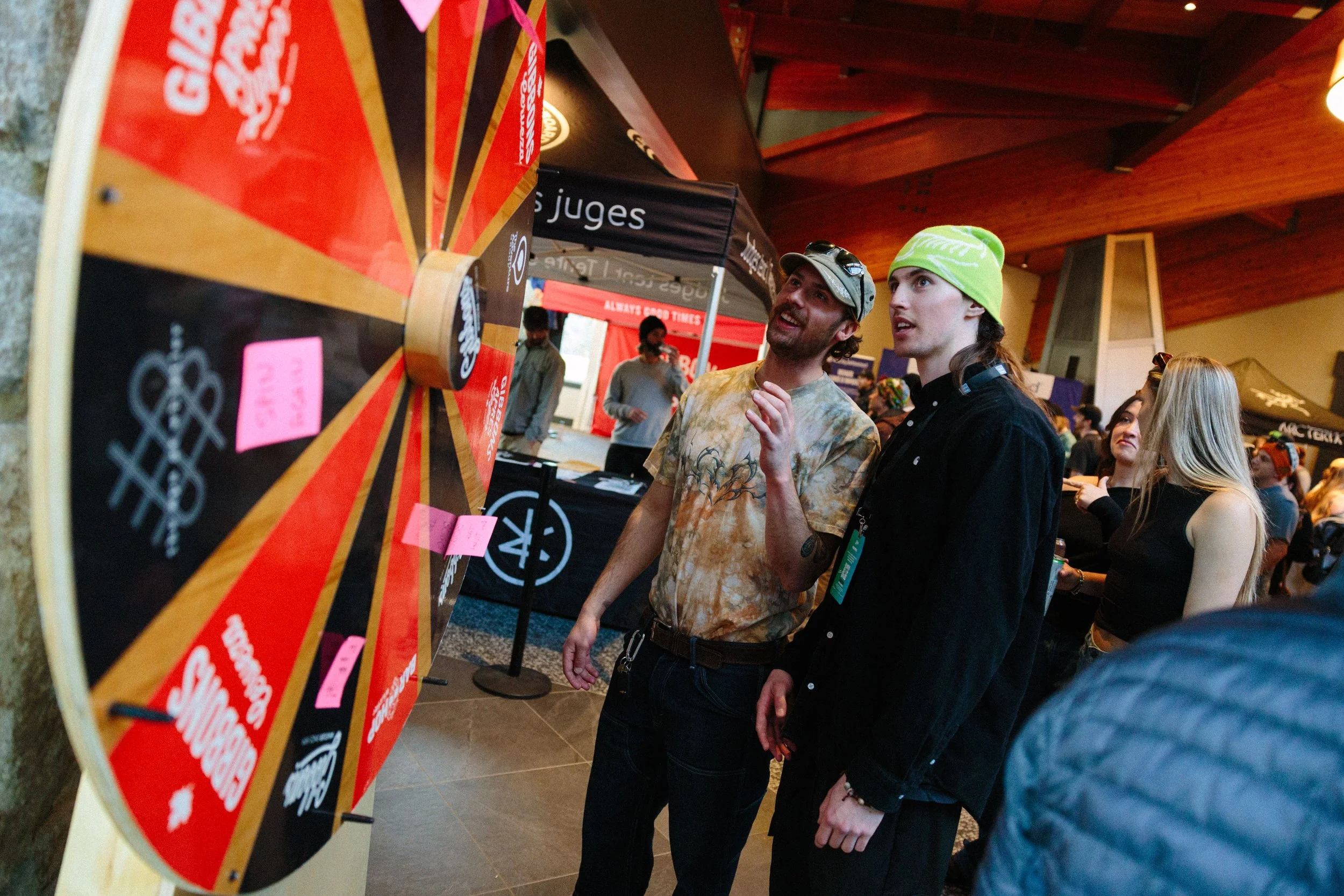 Two young men are at an event, looking at a prize wheel display. One is wearing a tie-dye shirt and baseball cap, the other has long hair, a green beanie, and a black shirt. People are visible in the background, some with drinks, inside a venue with 