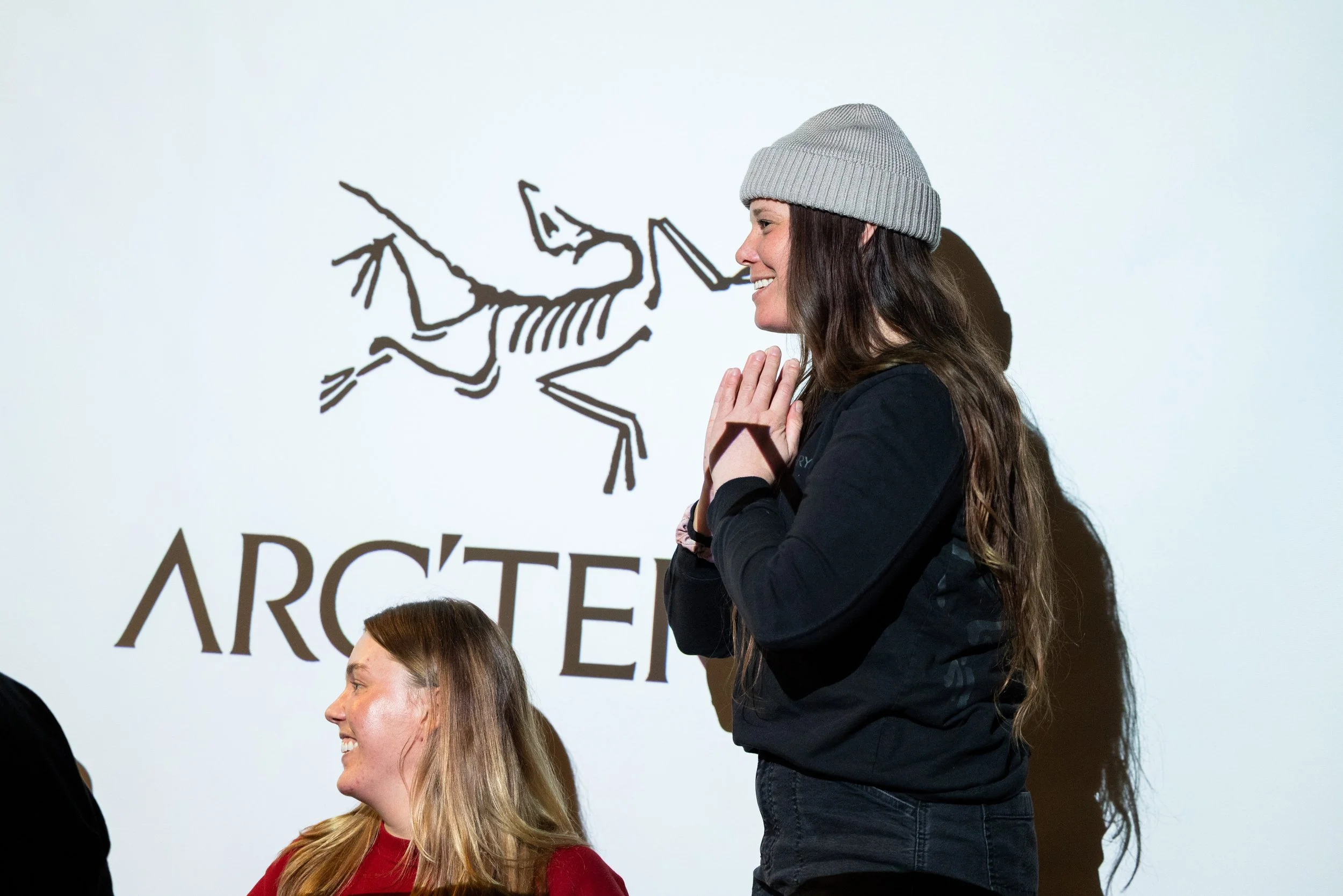 A woman with long brown hair wearing a gray beanie and black sweatshirt, standing with hands together in a prayer gesture, smiling in front of a wall with a drawing of a skeletal dinosaur and the word 'ARCHET'.