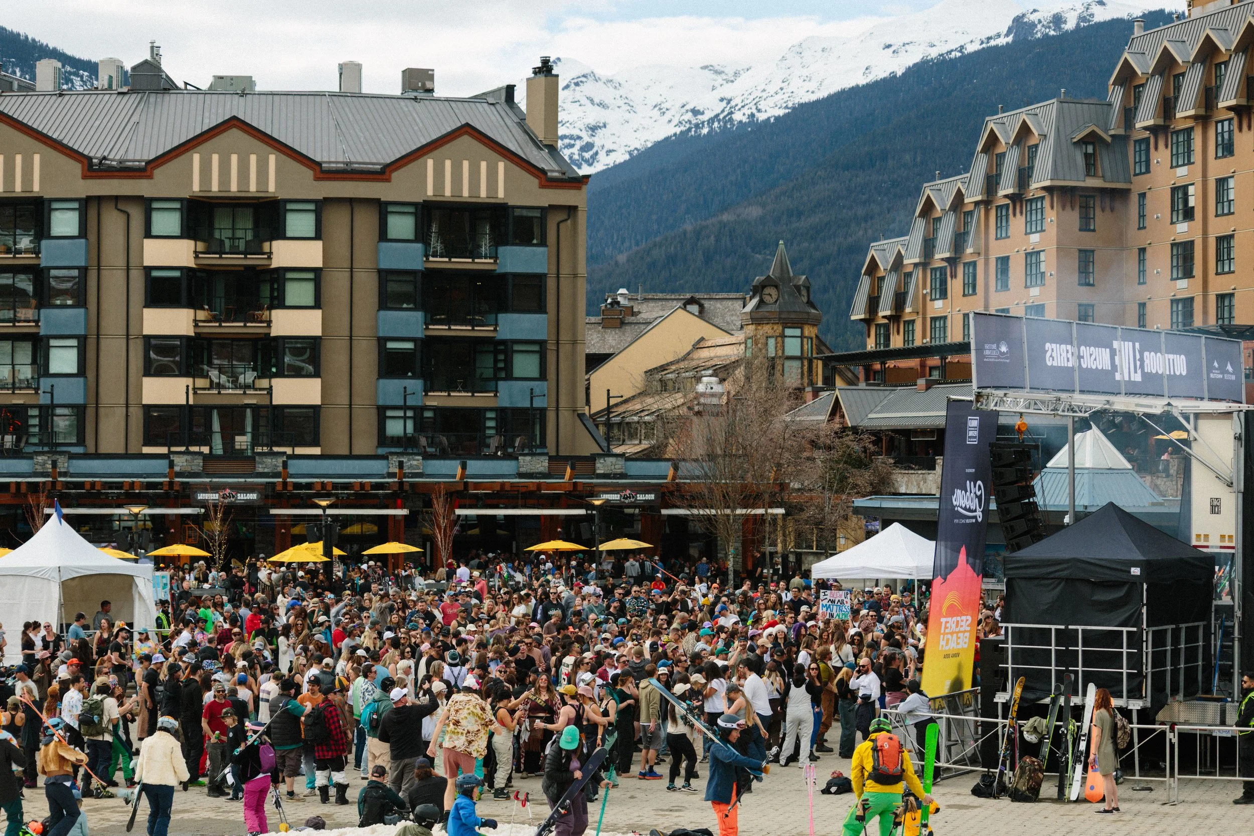 A large crowd gathered at an outdoor event in a mountain town, with tents, banners, and people walking around, surrounded by buildings with snow-capped mountains in the background.