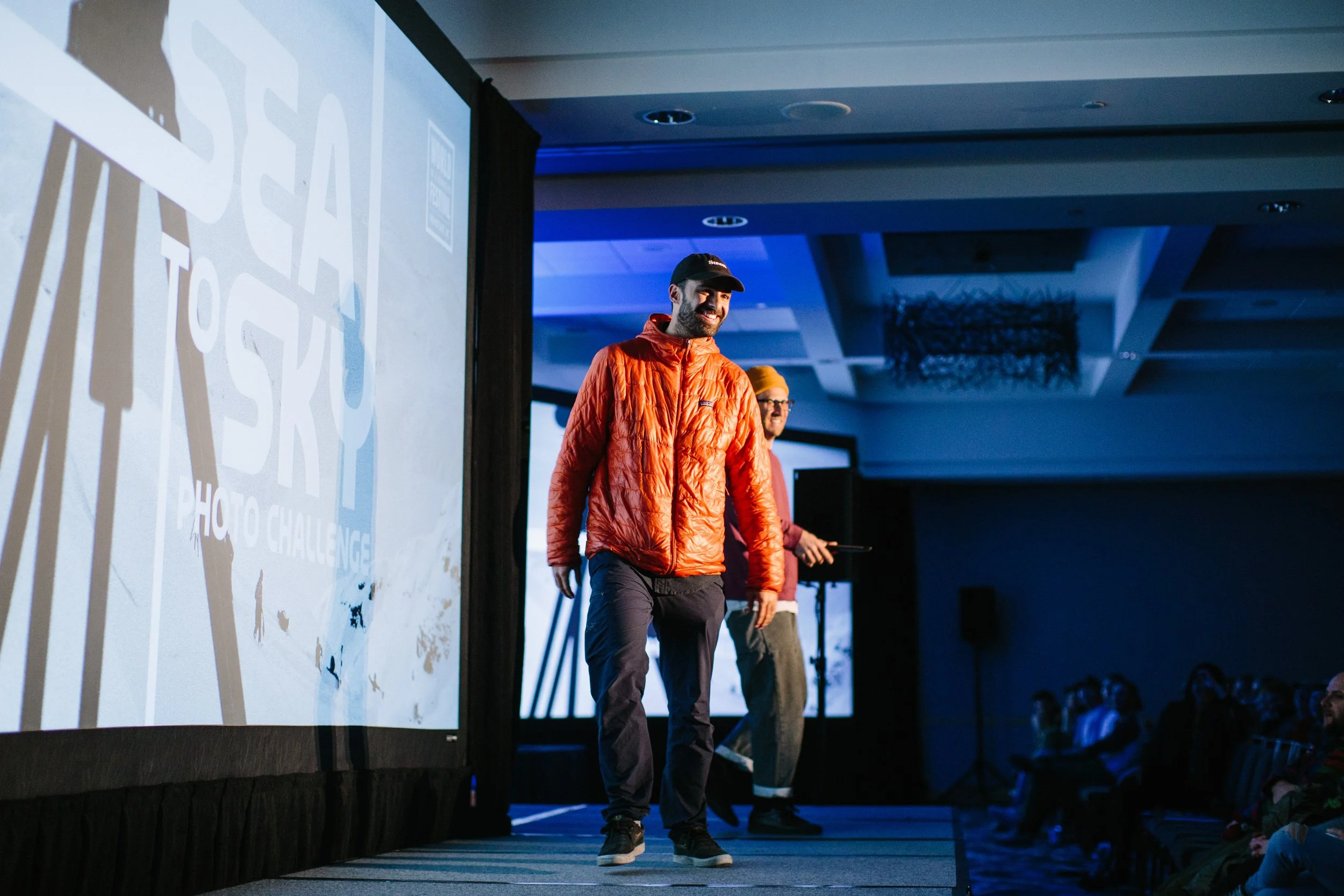 A man smiling on stage wearing an orange jacket and black cap at a photo challenge event, with a screen displaying the event's logo.