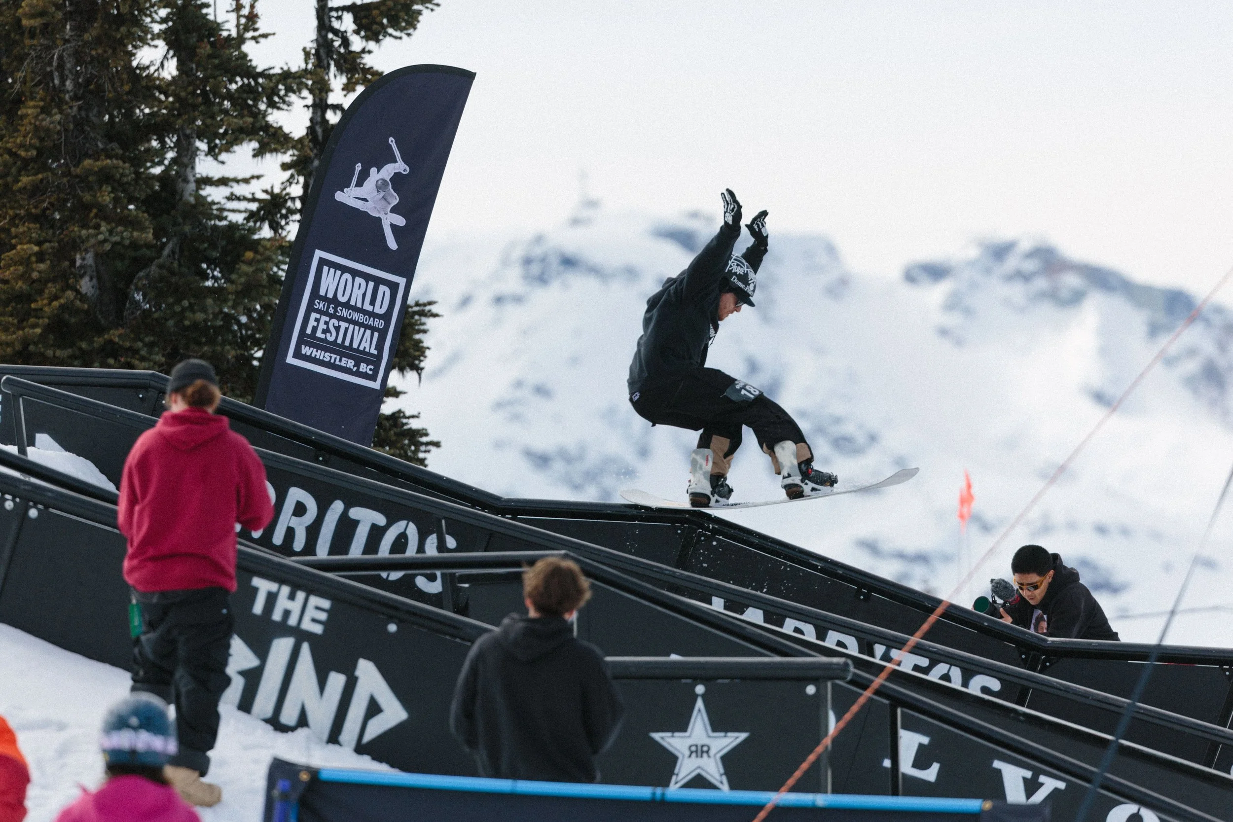 A skier mid-air performing a jump at the World Ski & Snowboard Festival in Whistler, BC, with some spectators watching nearby.