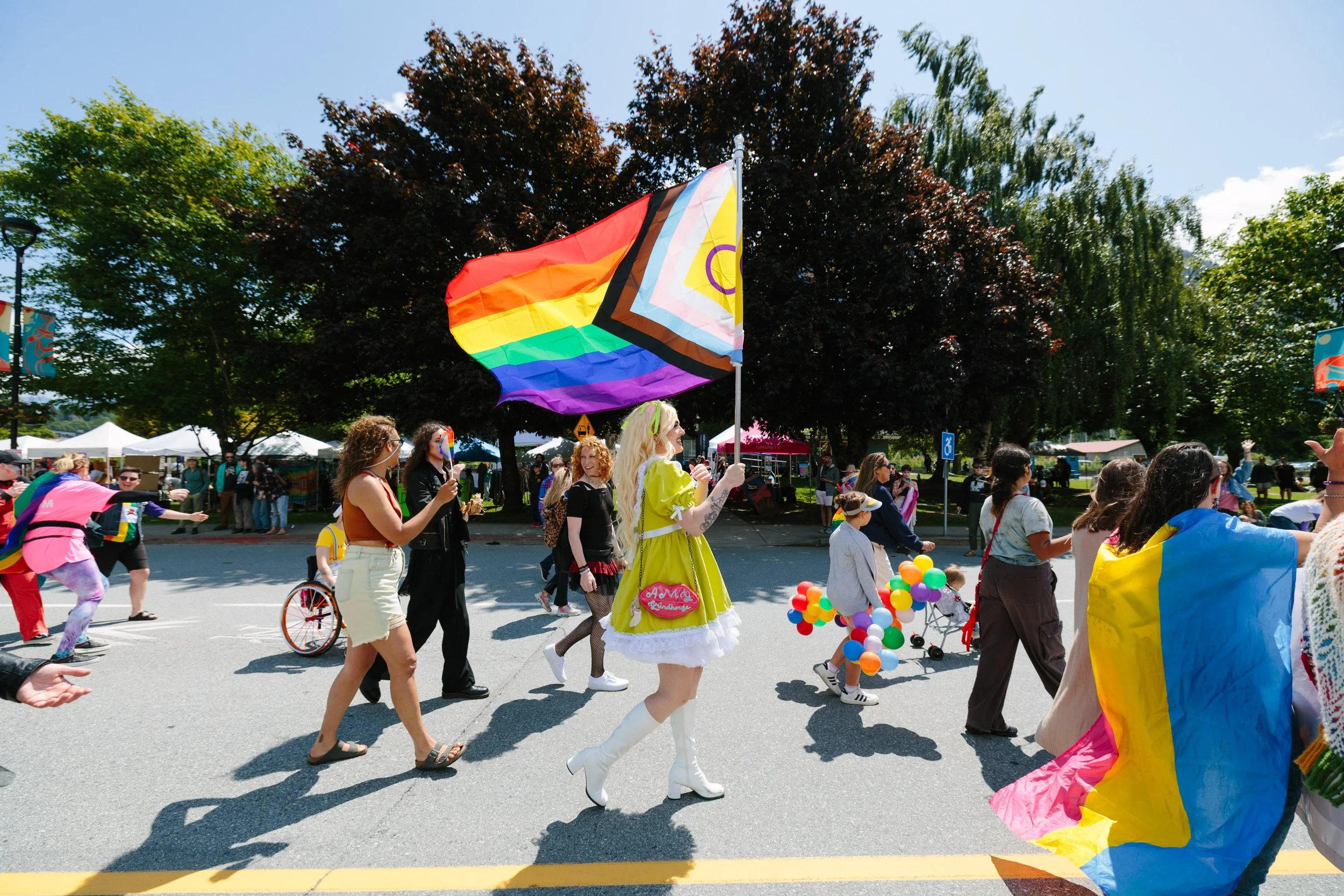 People participating in a parade during a pride celebration, carrying rainbow flags and colorful balloons, with tents and trees in the background on a sunny day.