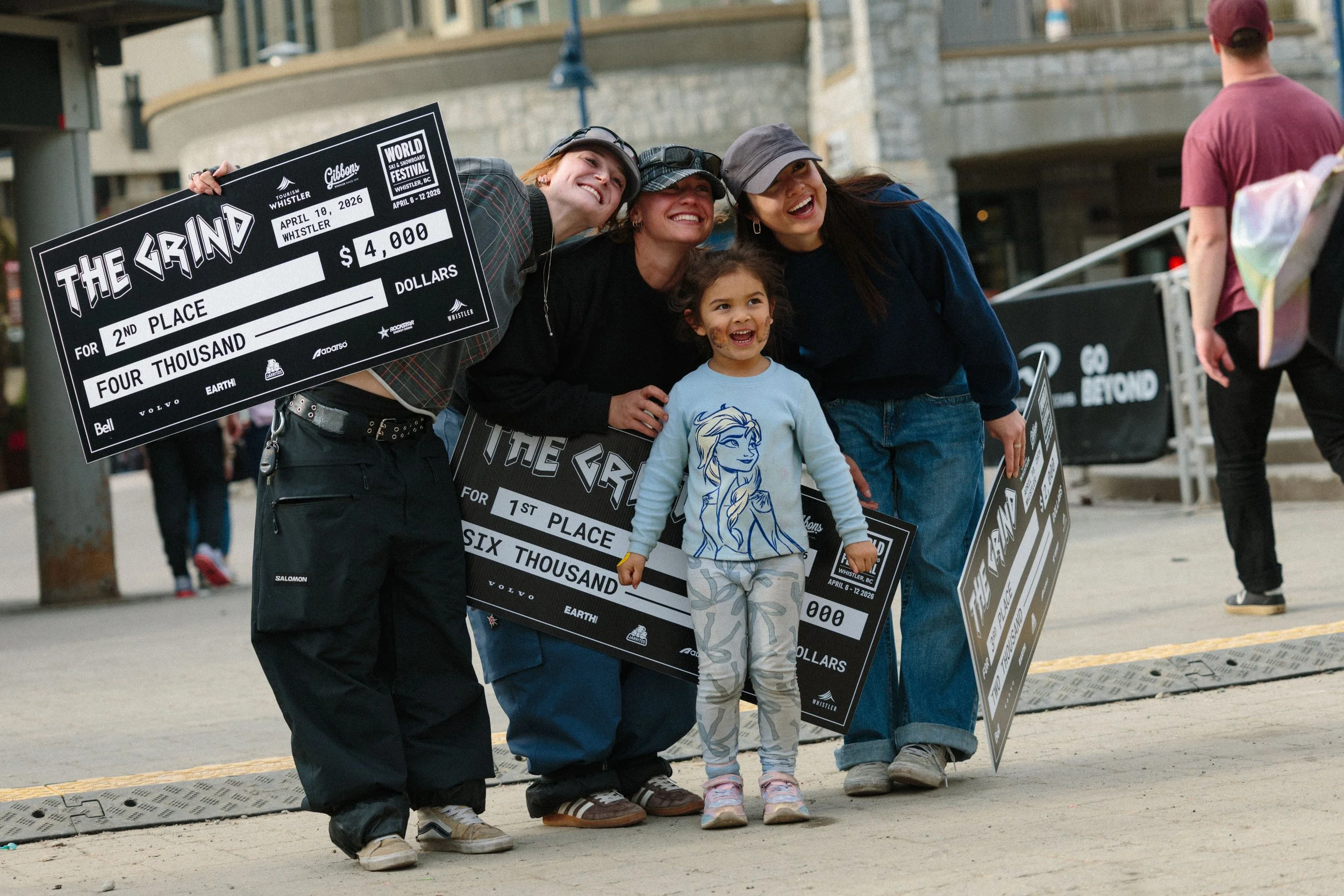 Group of four people, three women and one young girl, posing with a large black check for second place in a snowboarding event, outdoors in an urban area.
