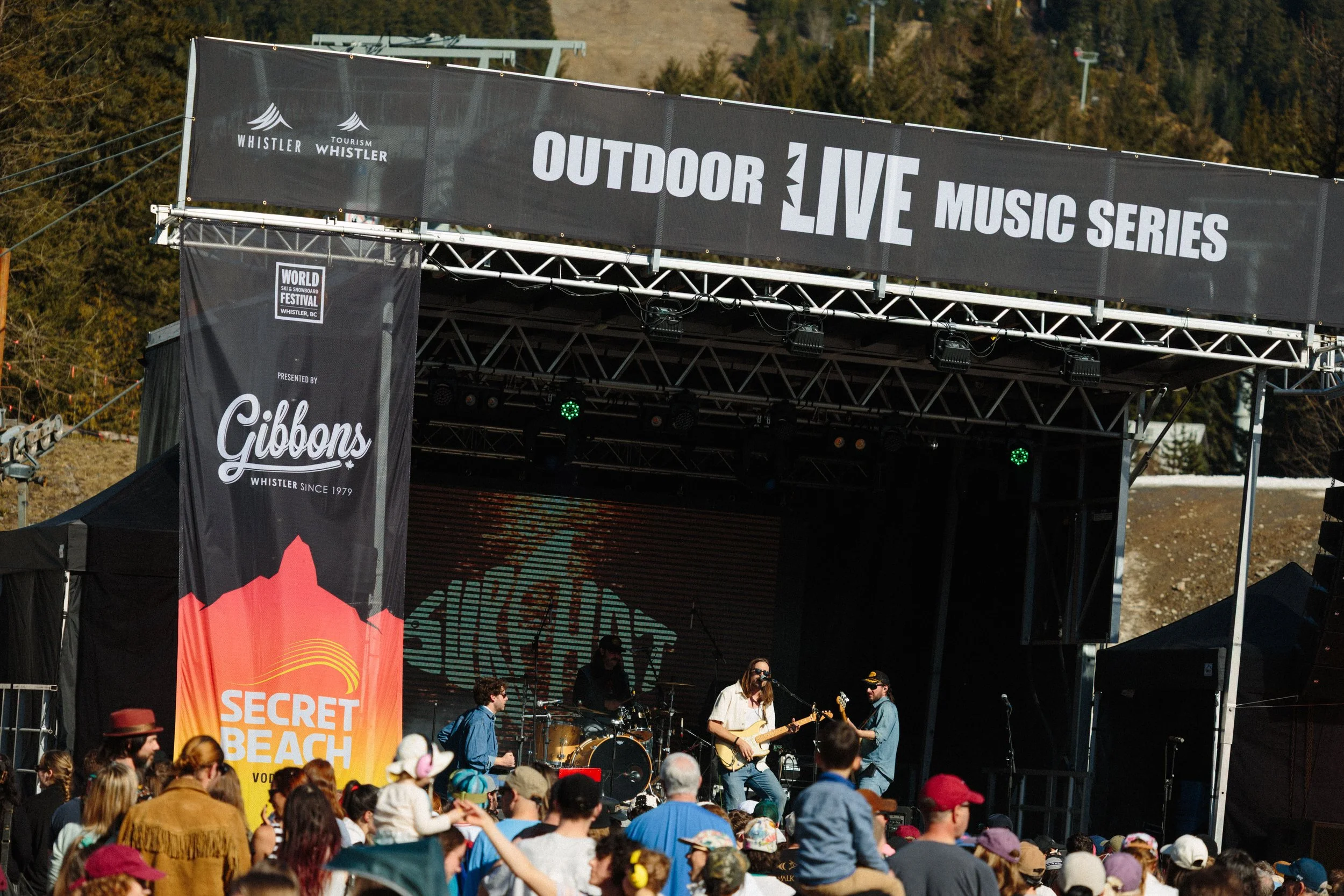 An outdoor live music performance on a stage at the Secret Beach festival in Whistler, Canada, with a crowd watching. The stage has banners, and there is a band playing instruments.