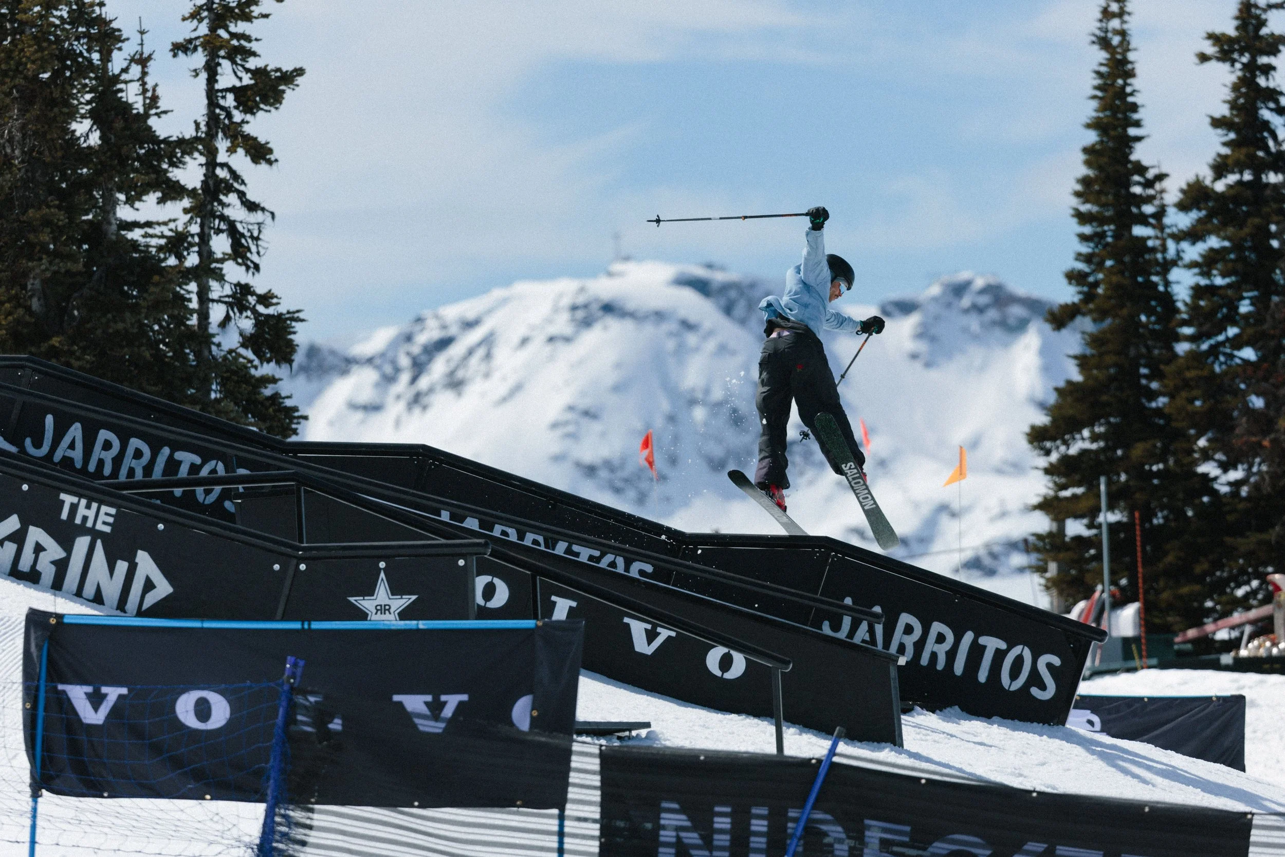 Skier in mid-air performing a trick at a winter outdoor ski park, with snow-covered mountains and pine trees in the background.