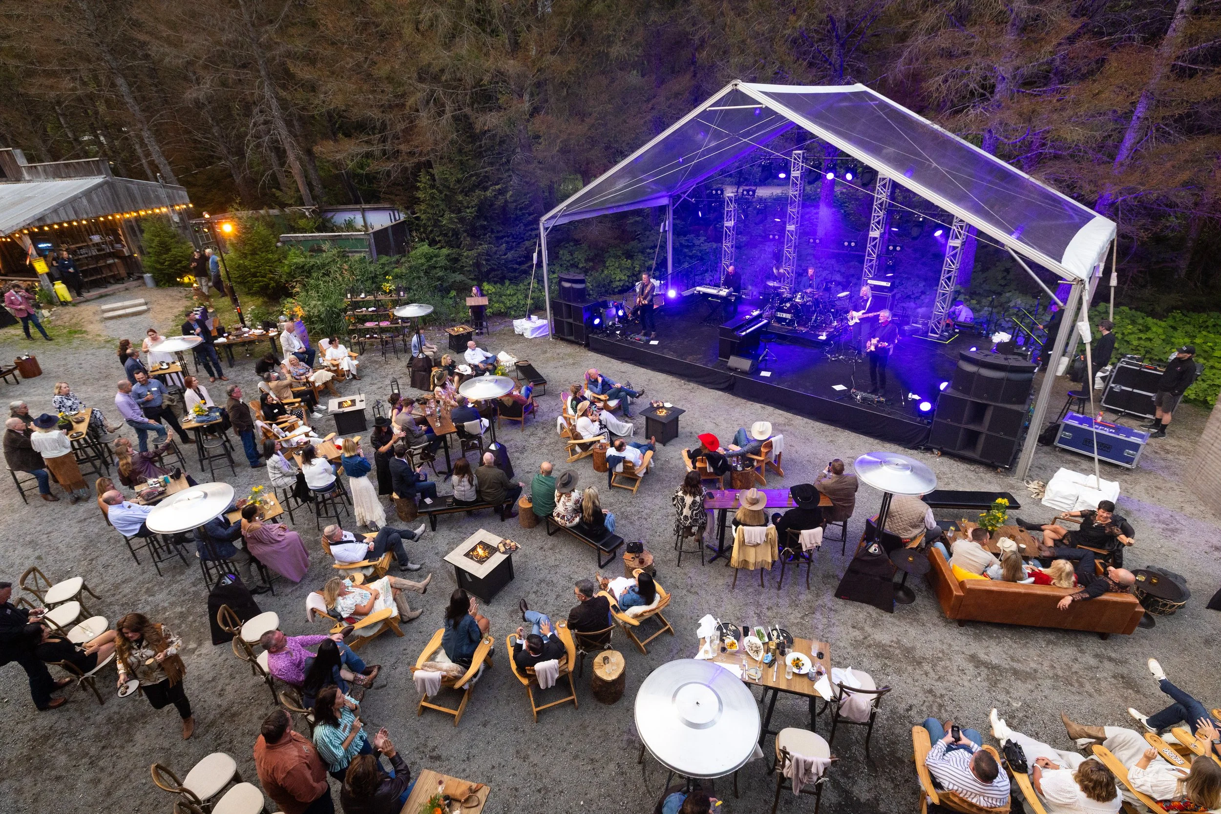 An outdoor concert scene with a band performing on a large stage under a canopy. Audience members are seated at tables and on benches, watching the performance in a yard surrounded by trees. Some tables have candles or small decorations. The scene is