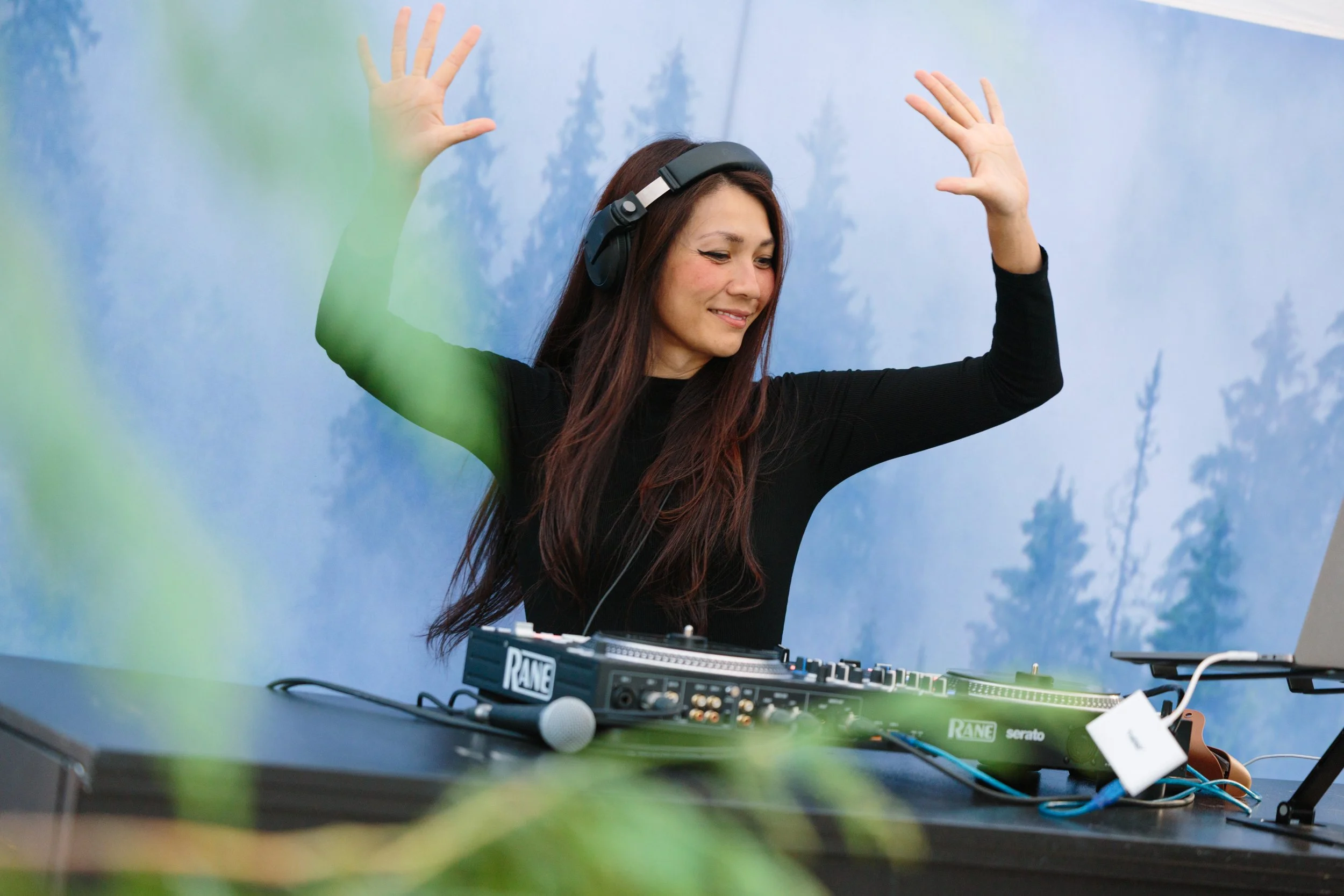 A woman with long brown hair, wearing a black long-sleeve shirt and headphones, is dancing and smiling behind DJ equipment with a background of a blue sky and trees.