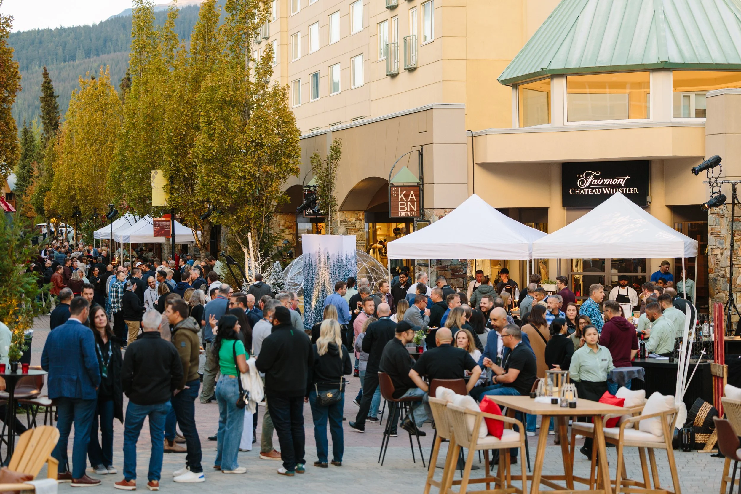 Outdoor gathering with many people socializing and dining at a street-side event in front of buildings with trees and mountains in the background.