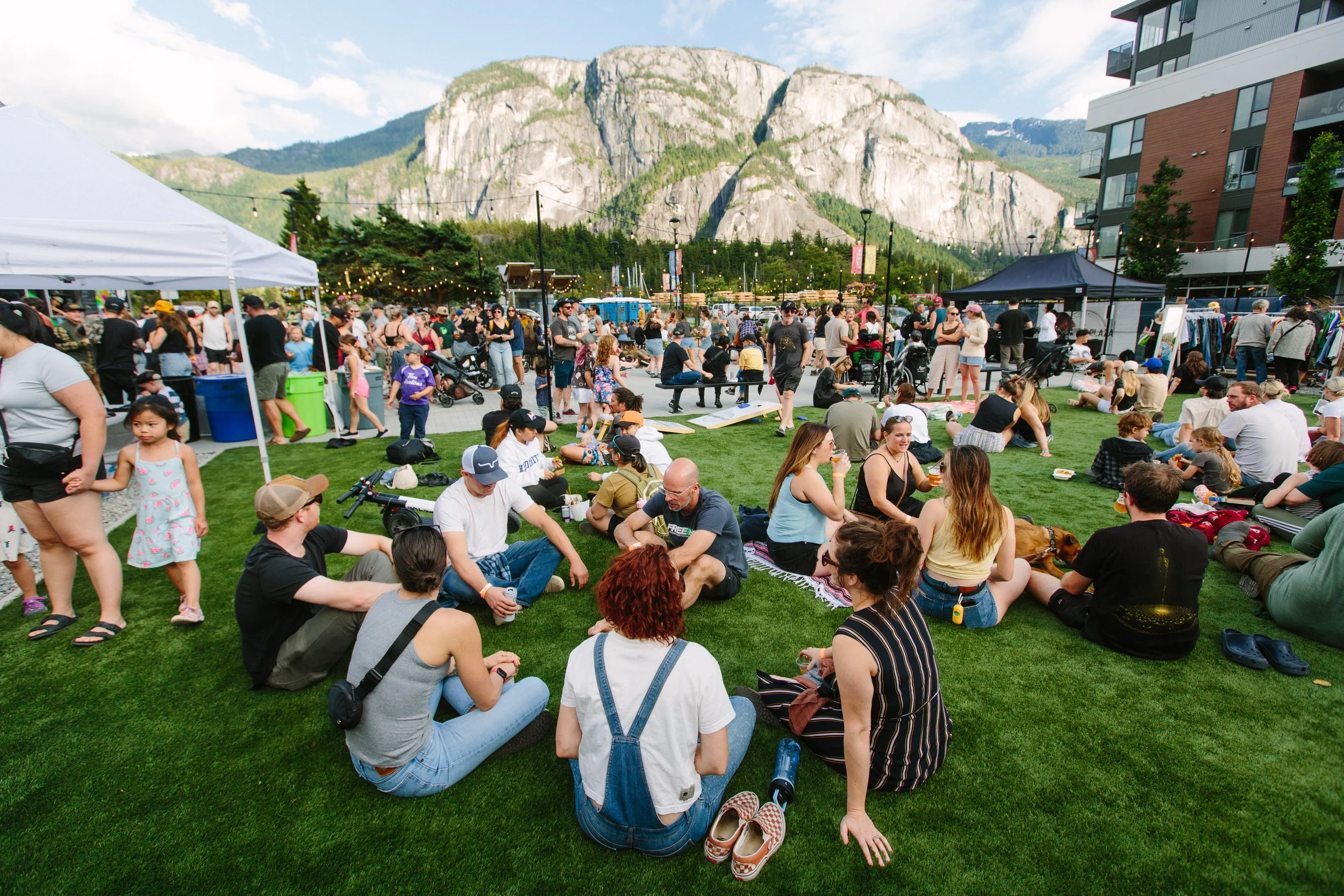 People gathered at an outdoor event during the daytime with mountains in the background. Some are sitting on grass while others are standing or walking around, with tents and vendor booths visible.