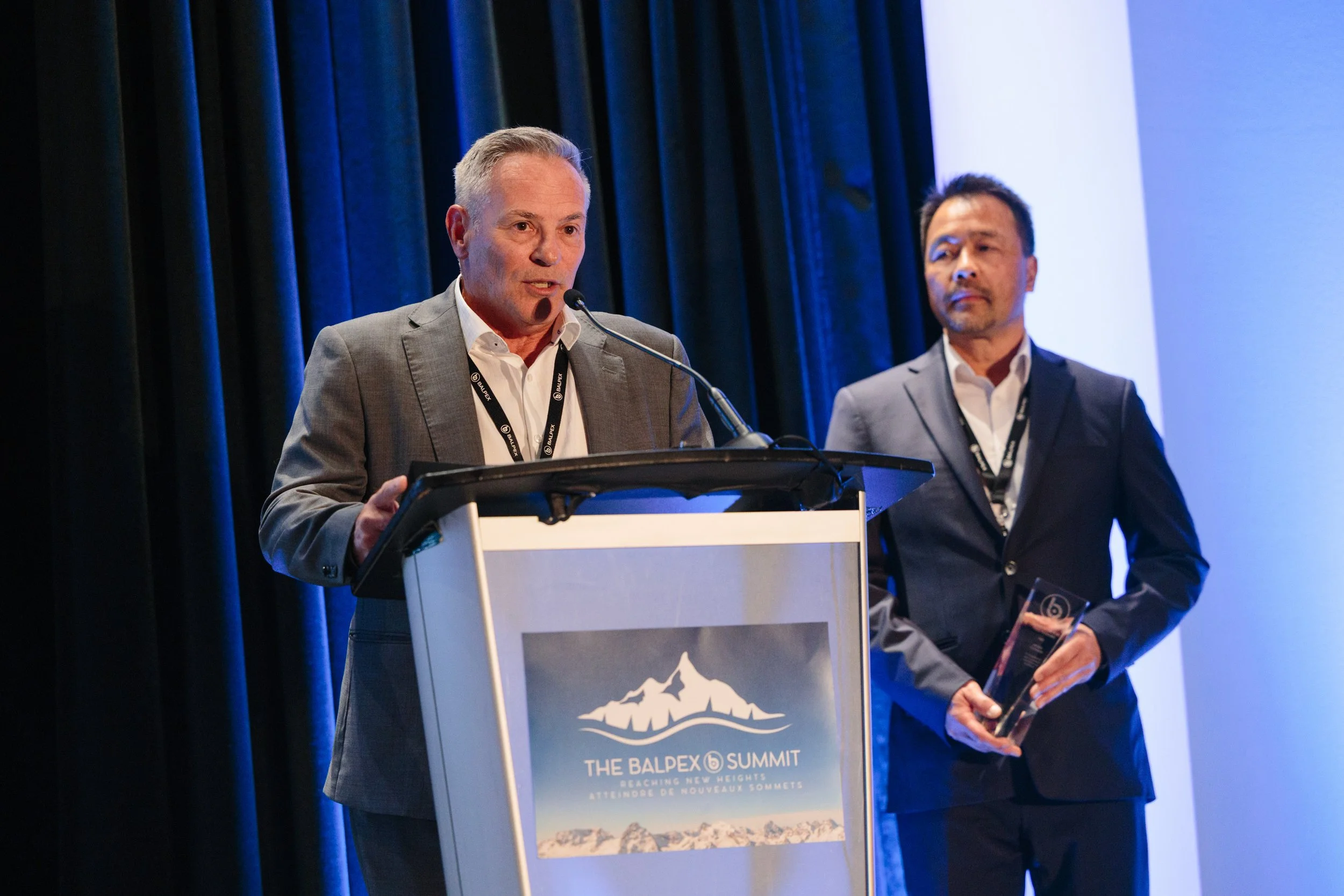 Two men in suits standing at a podium on a stage, one speaking into a microphone and the other holding an award, at the Balpex Summit.