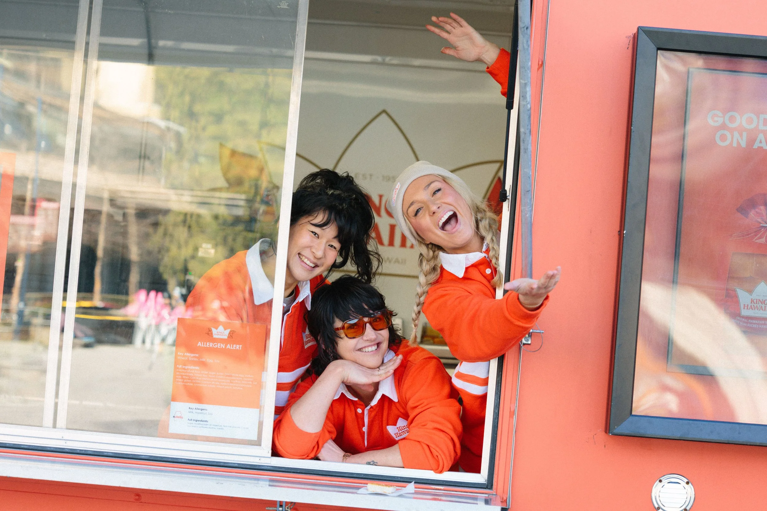 Four smiling women in orange uniforms, two with glasses, waving and looking out from a red food truck with a glass window and signs inside.