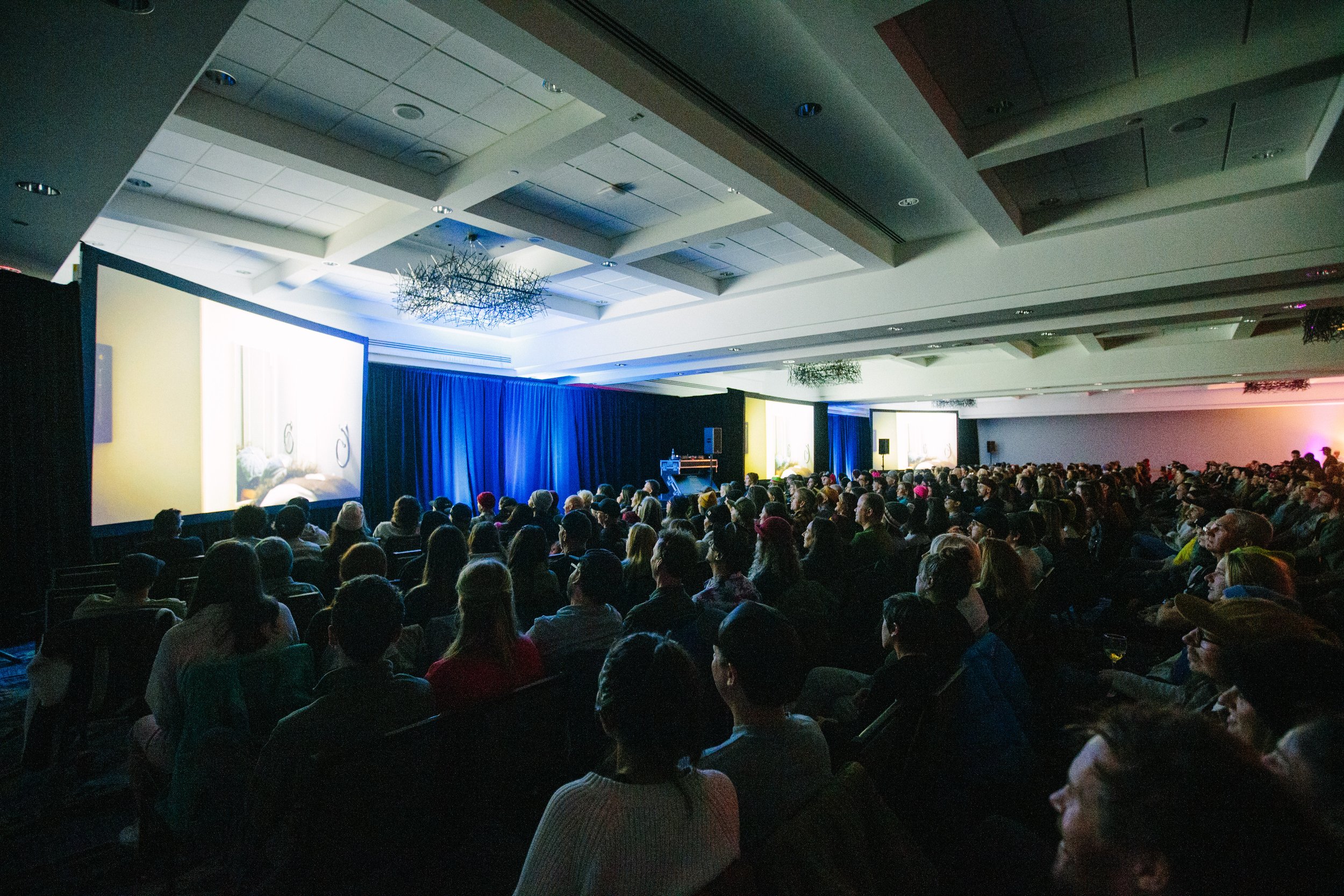 Large audience seated in a conference hall watching a presentation or film on two large screens.
