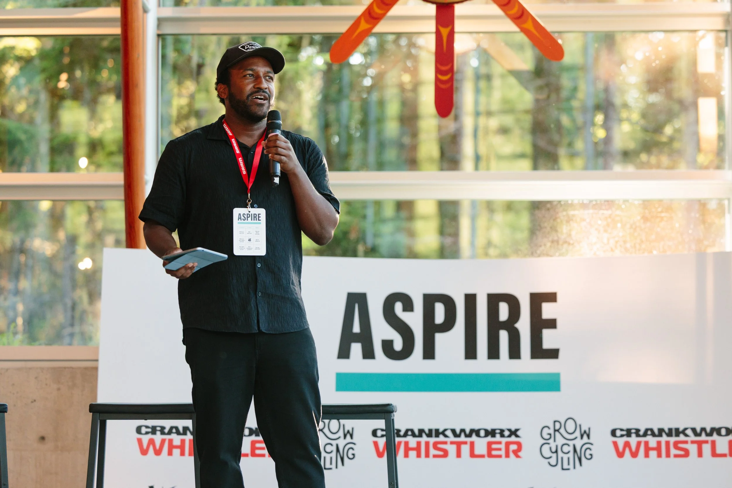 A man speaking into a microphone during a presentation at an event called ASPIRE, with a large ASPIRE sign behind him and a blurred background of trees and windows.