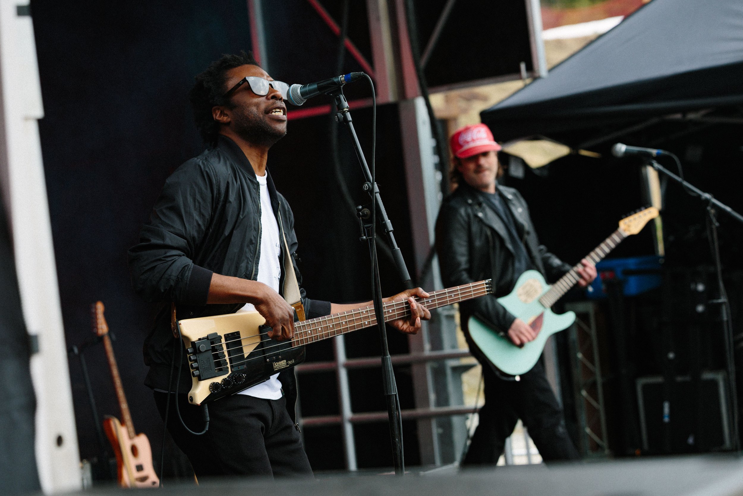 Two musicians performing on stage, one with sunglasses playing a bass guitar and the other in a red cap playing an acoustic guitar.