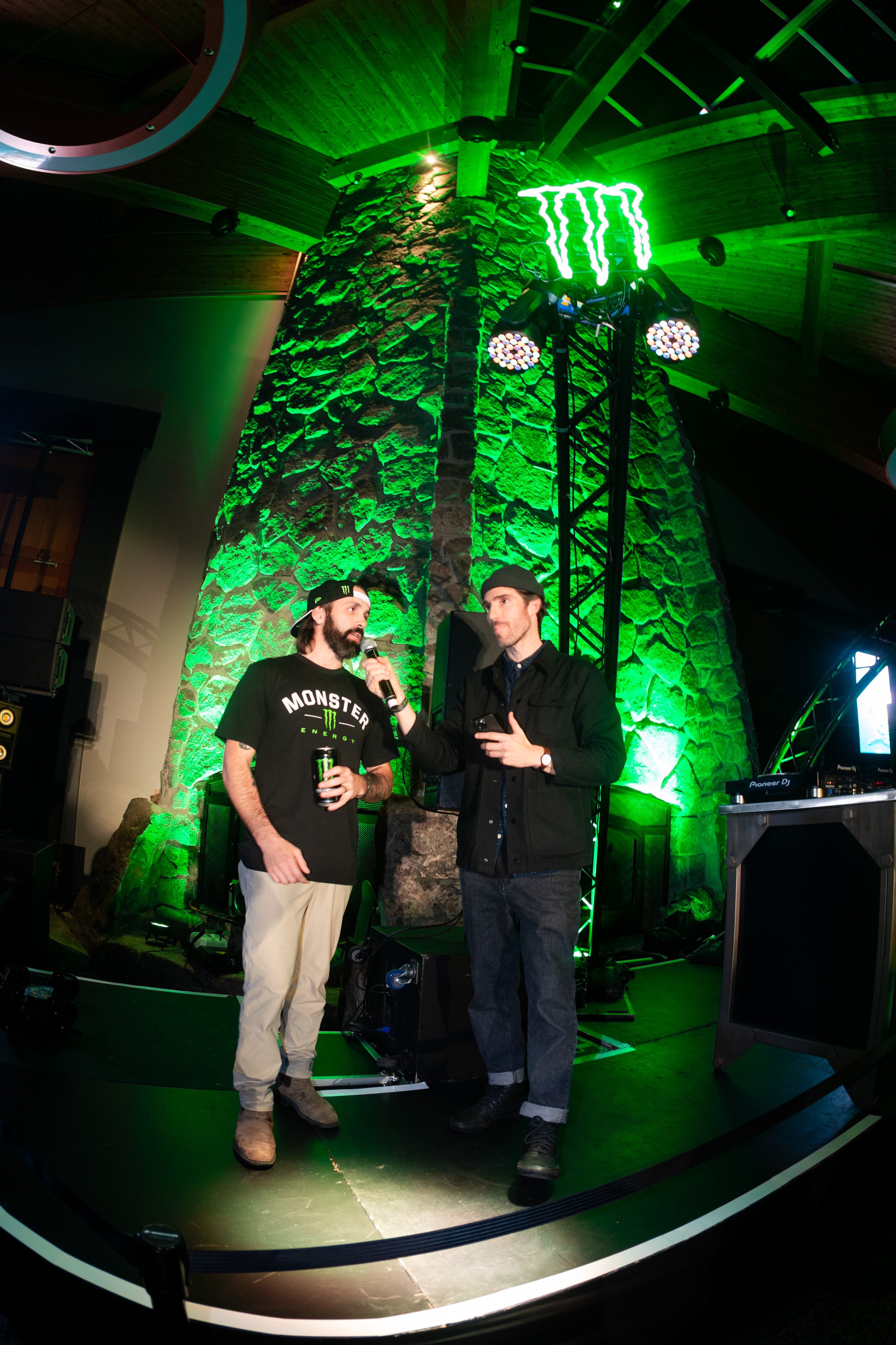Two men on a stage with green lighting, one holding a microphone and wearing a black Monster Energy t-shirt, the other holding a phone, standing in front of a large stone chimney with a neon Monster Energy logo overhead.