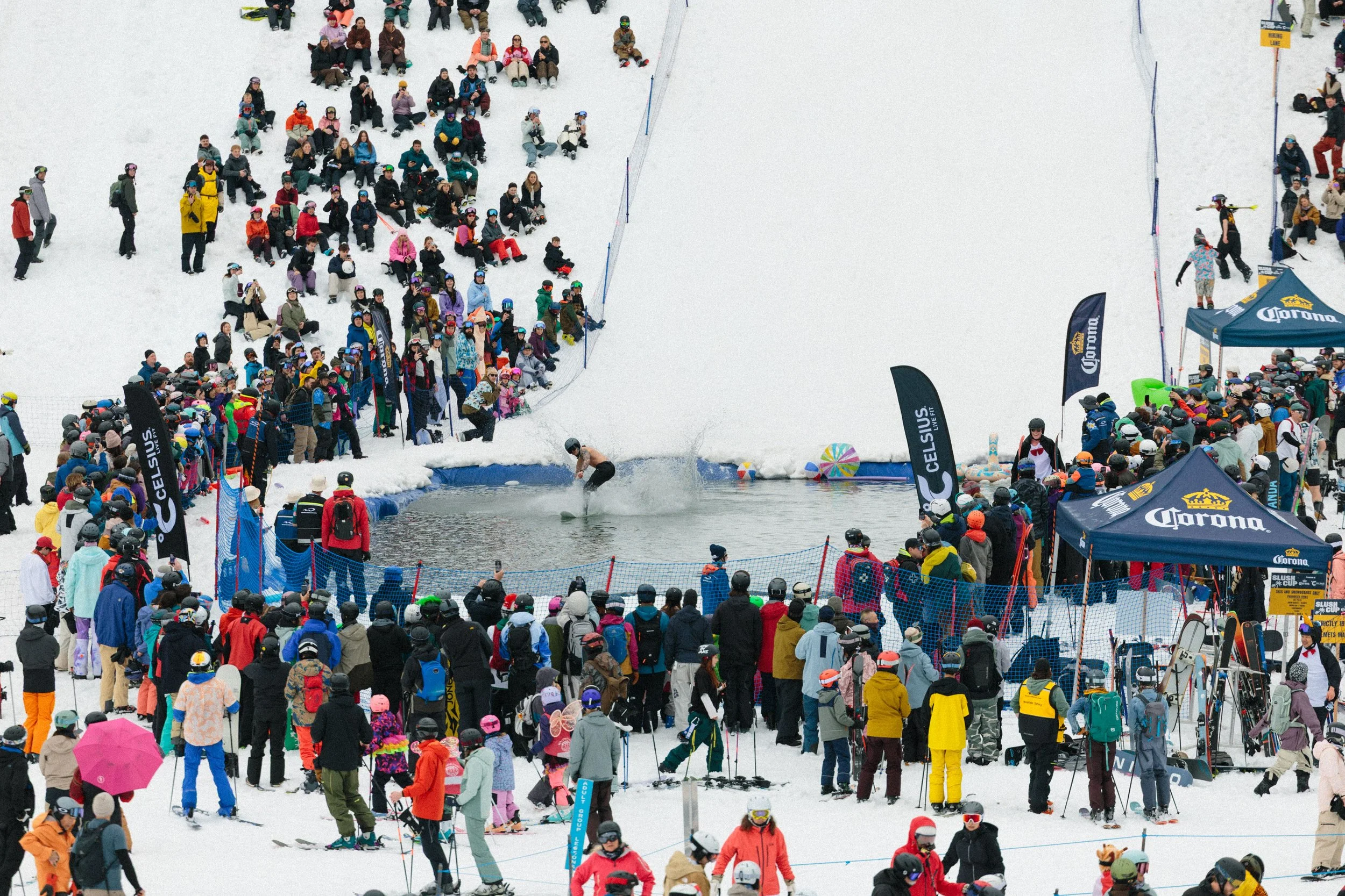 A large crowd of people gathered at a snowy venue for a winter event. Many are wearing colorful winter clothing and helmets. There is a water descent with a skier or snowboarder in the water, and there are flags and tents nearby, indicating an organi