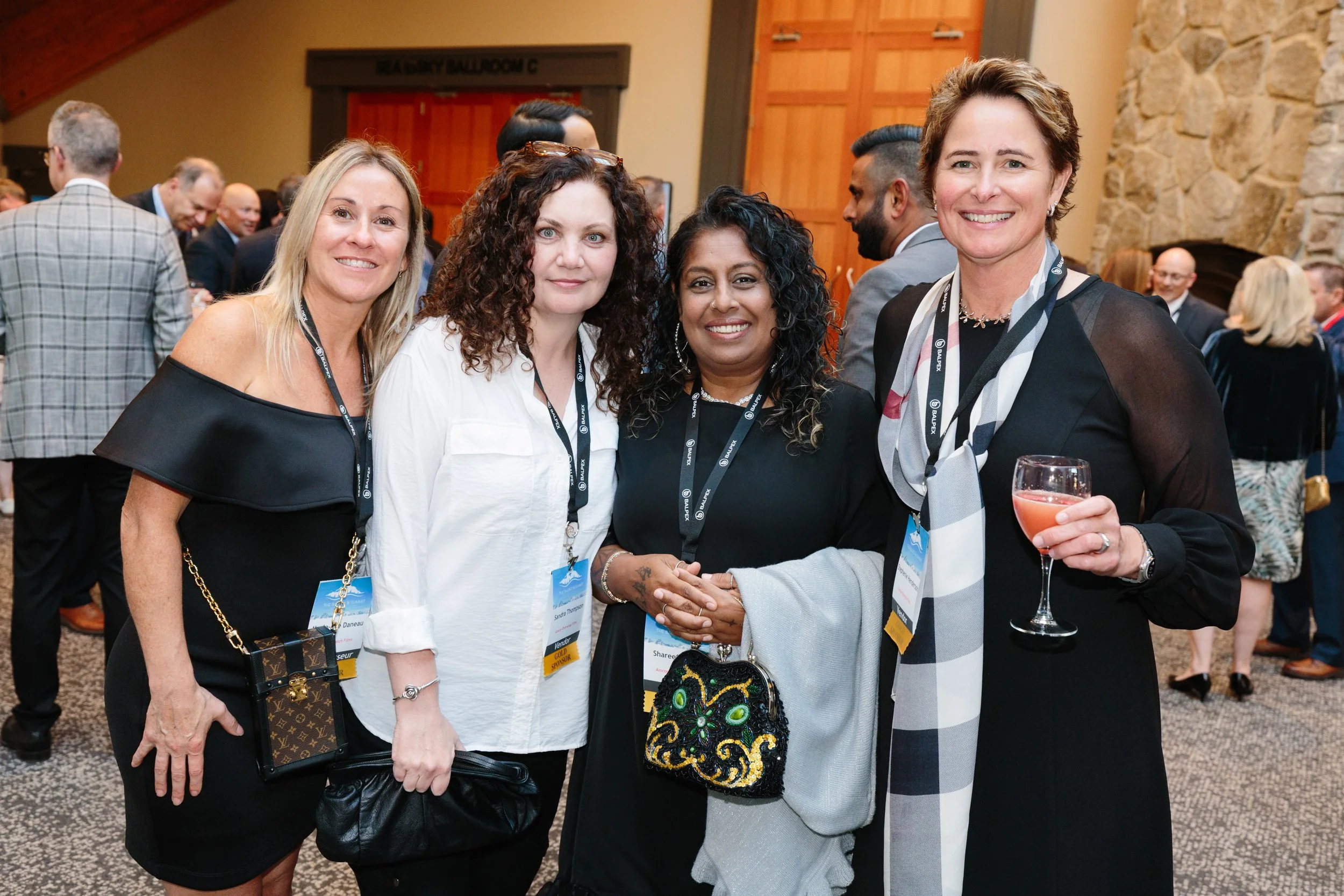 Four women smiling and standing together at a professional event, with one holding a glass of pink drink. They are wearing name tags and are surrounded by other attendees in a conference room with stone and wood decor.