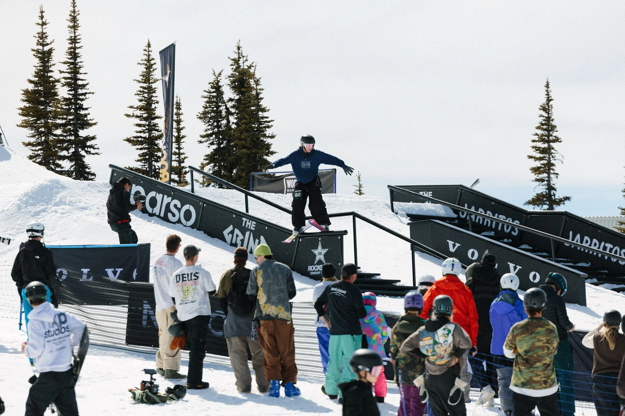 A snowboarder performs a trick on a rail at a snowboarding event, with spectators watching nearby on a snowy slope under a cloudy sky, surrounded by evergreen trees.