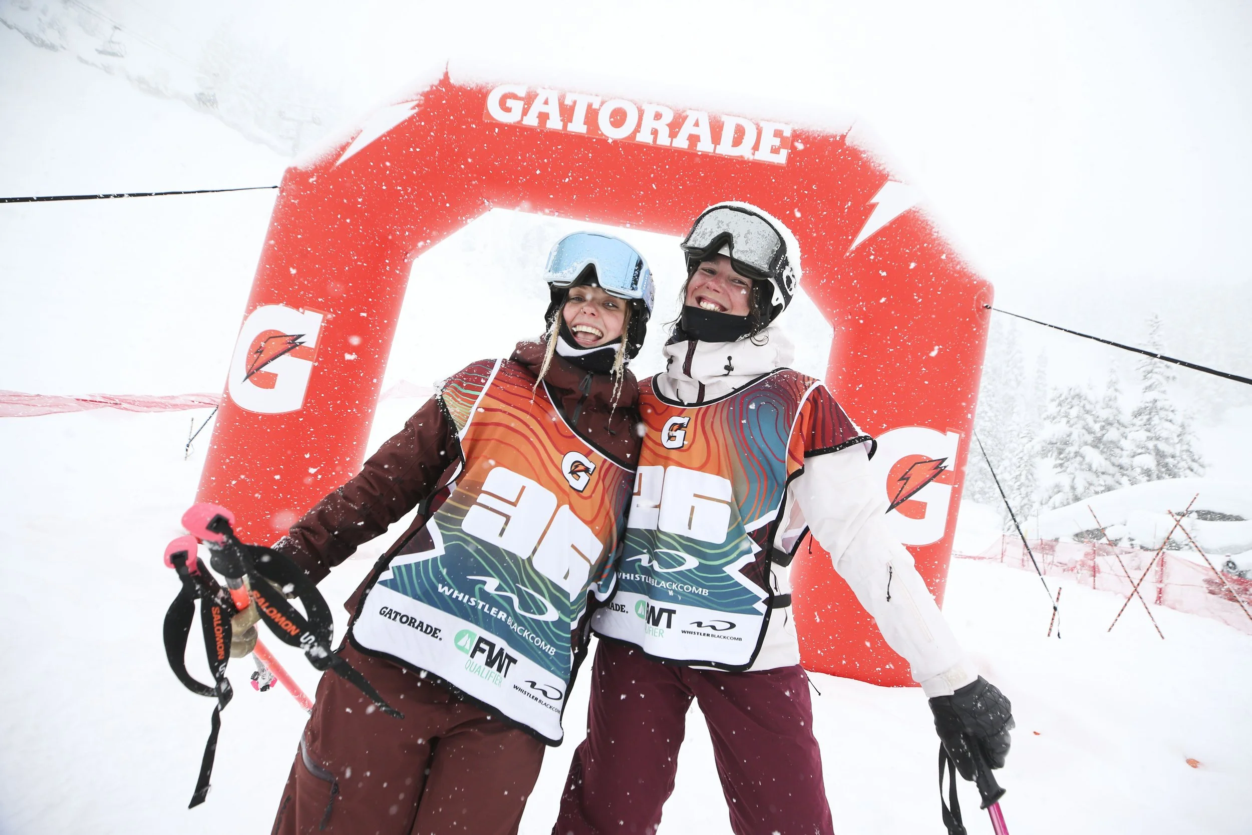 Two smiling snowboarders standing under a red Gatorade arch in a snowy, foggy mountain setting, wearing helmets, goggles, and colorful snowboarding gear.