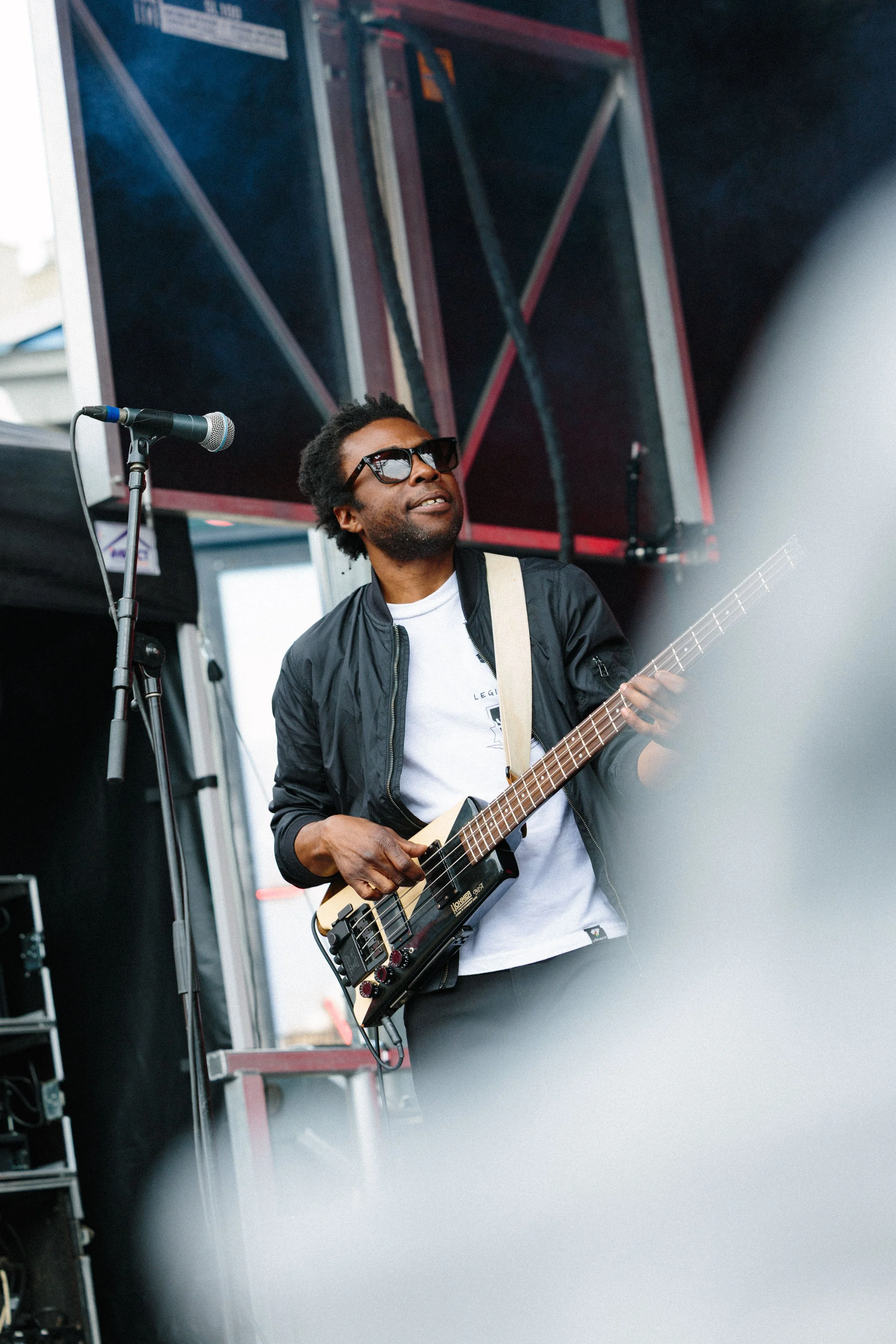 A man wearing sunglasses playing an electric guitar on a stage with a microphone in front of him and speaker cabinets in the background.