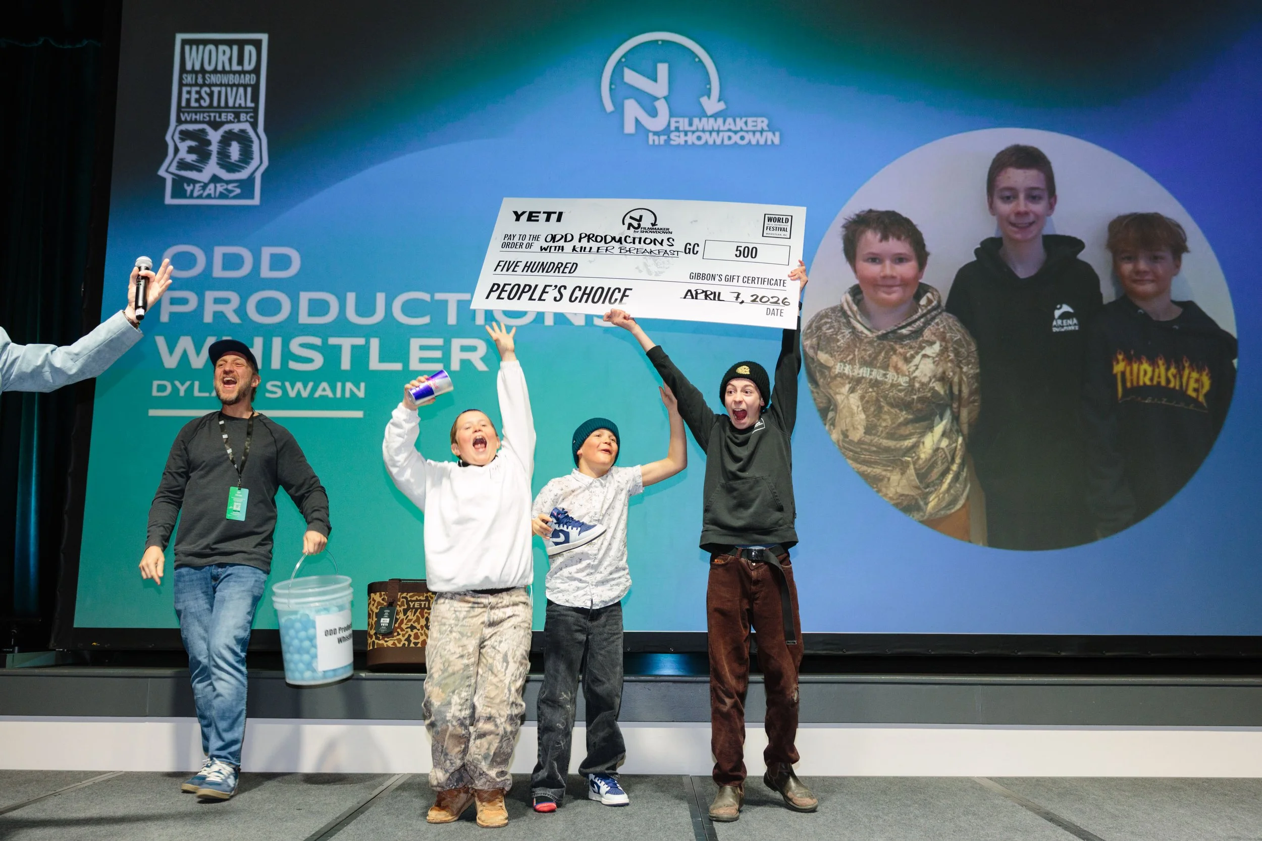 A group of children and adults on stage celebrating at the World Ski & Snowboard Festival in Whistler, British Columbia. One child holds a large ceremonial check for 500 dollars made out to Qard Productions.