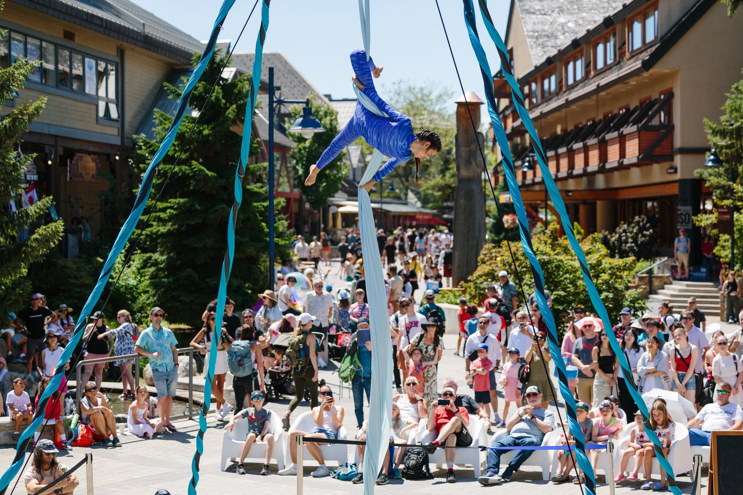 Aerial silk performer in a blue outfit performing on blue silks in an outdoor shopping district, with a large crowd watching.