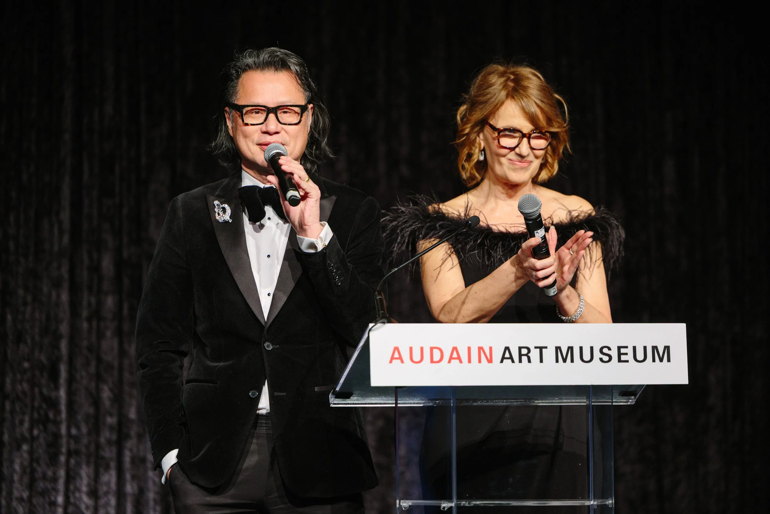 Two people speaking at a podium with a sign reading 'AUDAIN ART MUSEUM'. The man on the left is wearing a black tuxedo with a bow tie, and the woman on the right is wearing a black dress with feathered shoulders and glasses.