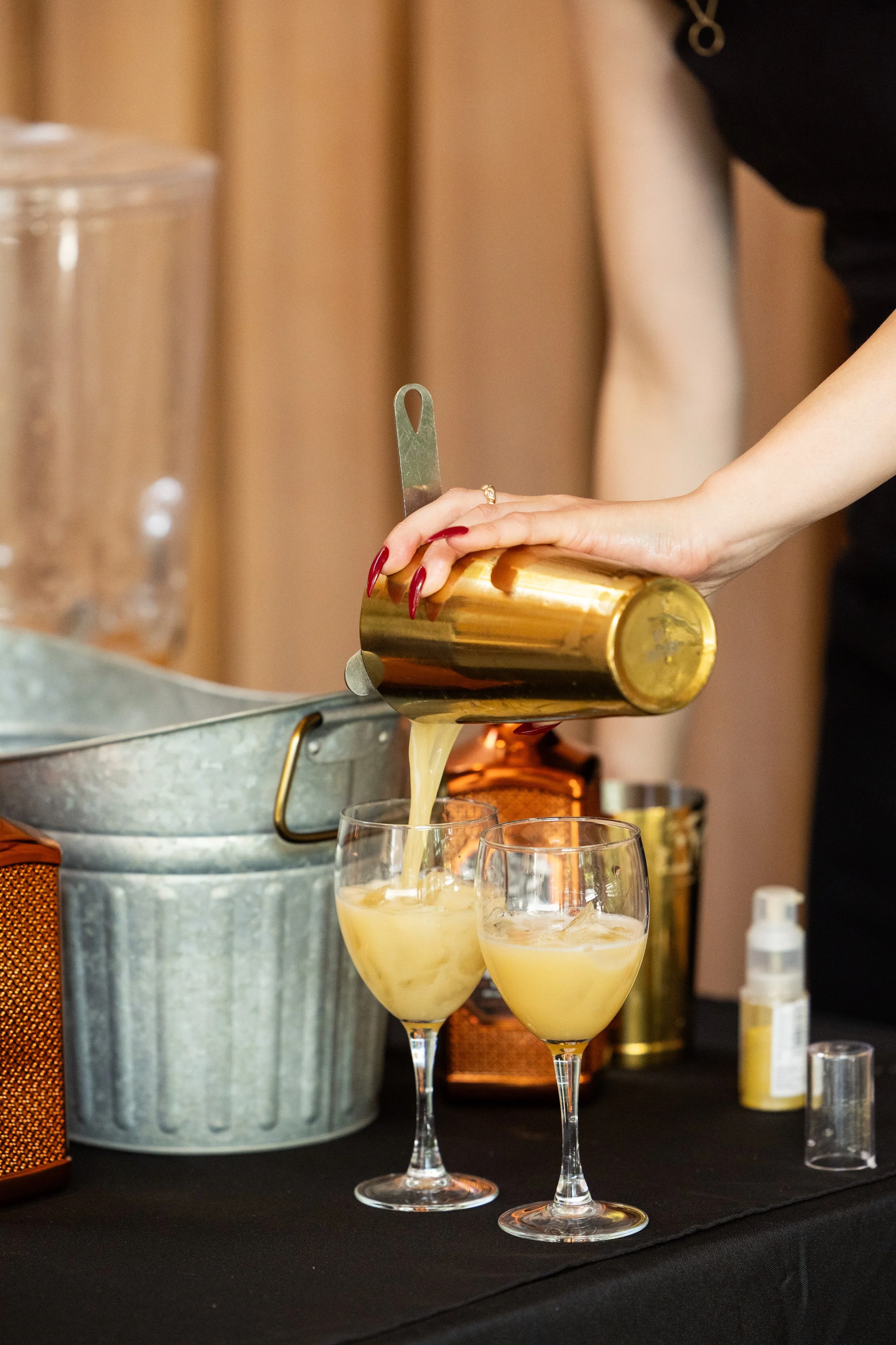 Person pouring a yellow cocktail from a gold shaker into two wine glasses on a black table.